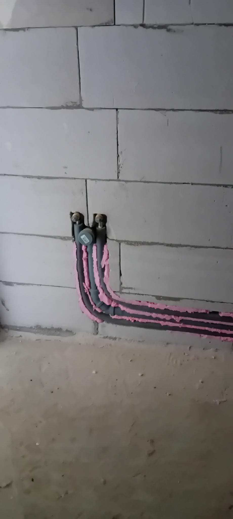 Szare rury instalacji hydraulicznej, zabezpieczone różową pianką, prowadzone wzdłuż ściany z białych bloczków. Widoczne przyłącza.