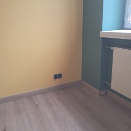 Bartlomiej kudelko - Pomalowane na żółto ściany z gniazdkami, drewniana podłoga i fragment ściany pomalowanej na zielono z grzejnikiem. Widok na róg pokoju po remoncie.
