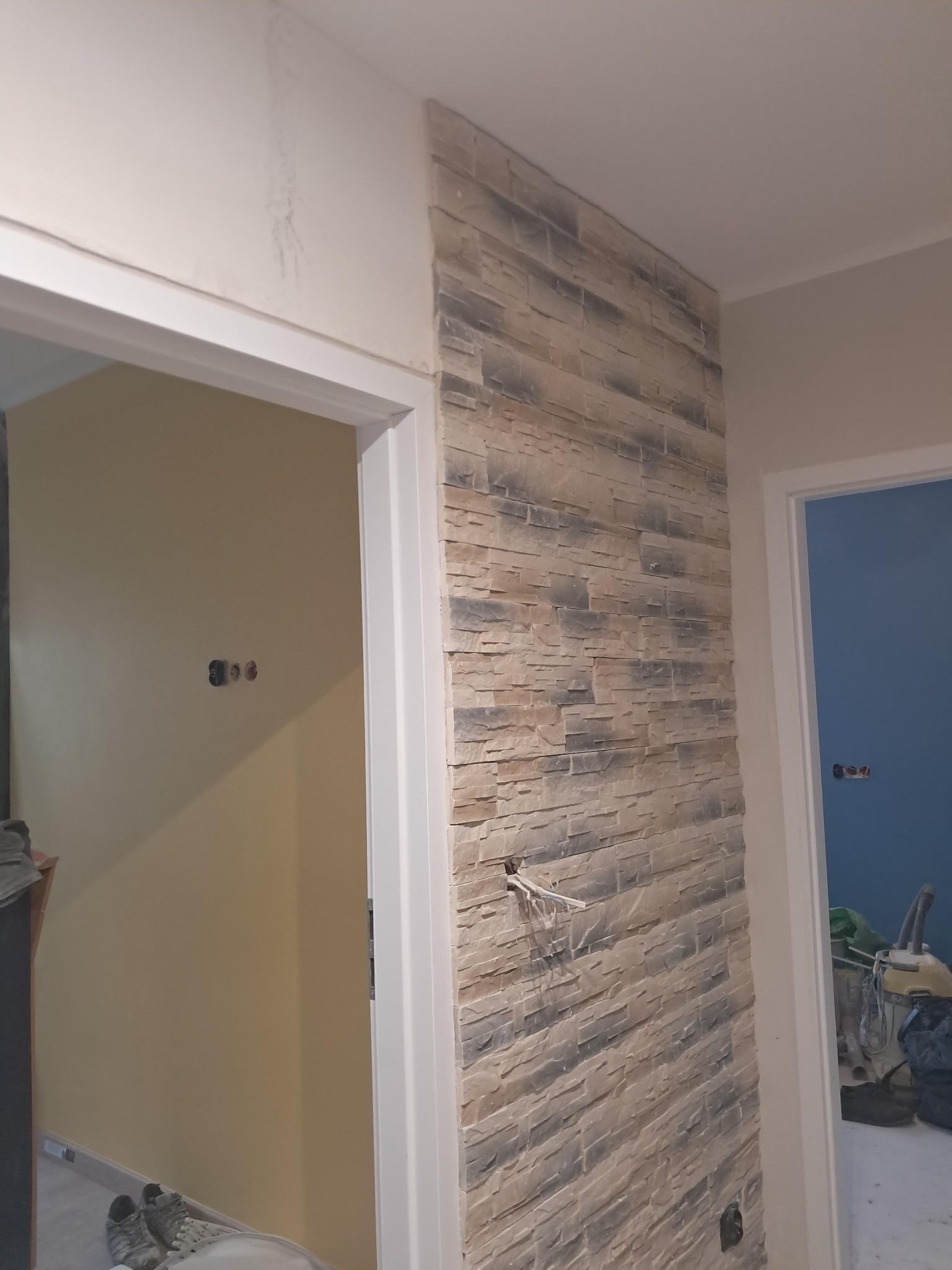Wykończenie ściany imitacją kamienia w korytarzu, widoczne gniazdka elektryczne i fragmenty drzwi do sąsiednich pomieszczeń. Dekoracyjny element wykończenia wnętrza.