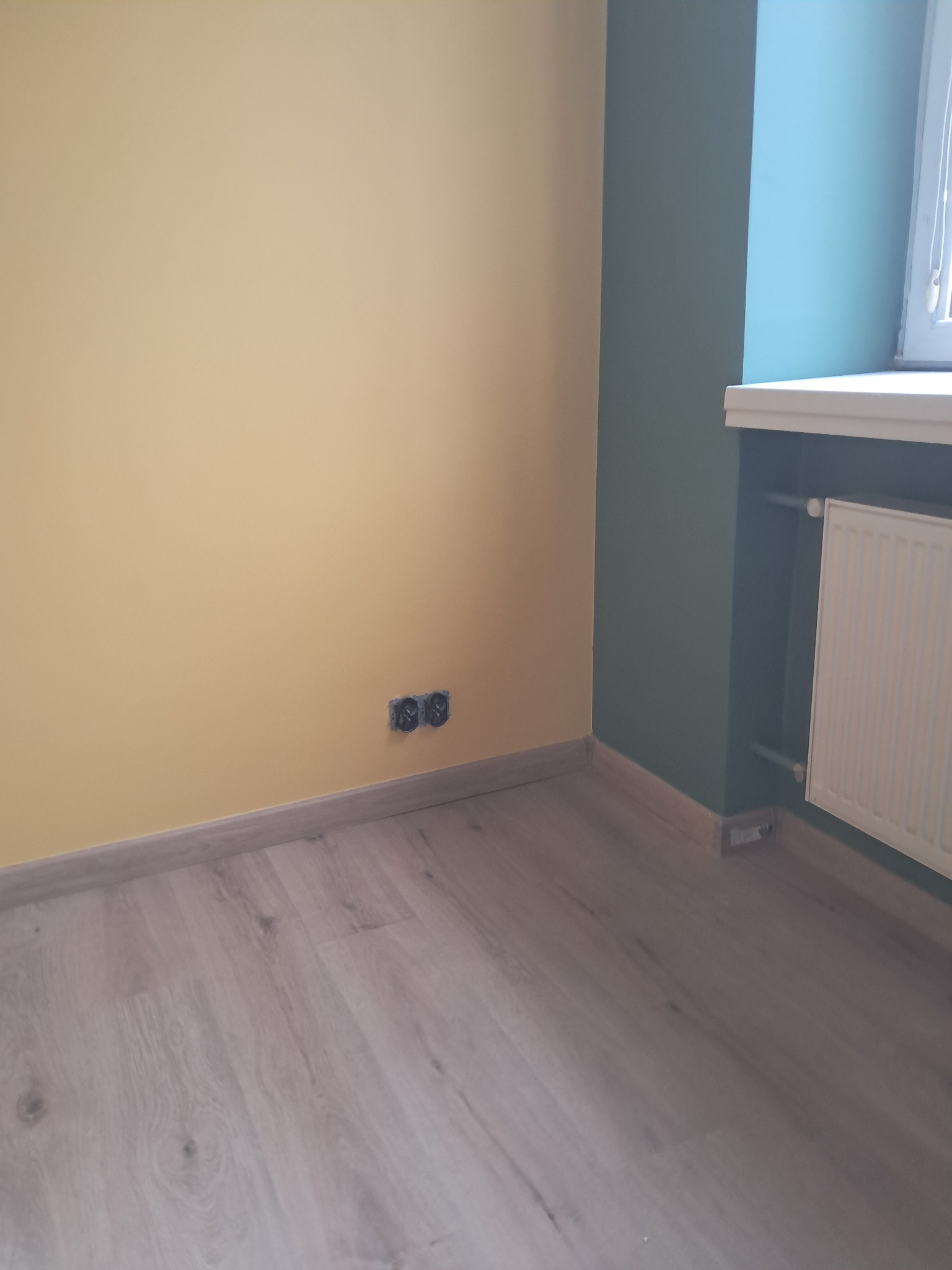 Pomalowane na żółto ściany z gniazdkami, drewniana podłoga i fragment ściany pomalowanej na zielono z grzejnikiem. Widok na róg pokoju po remoncie.