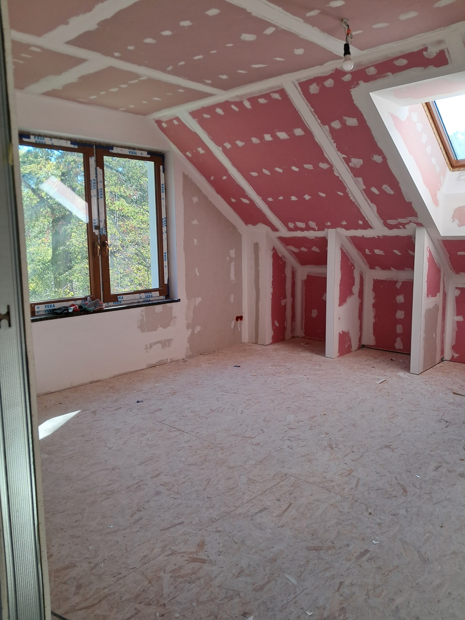 Poddasze w trakcie adaptacji: płyty gipsowo-kartonowe na skosach i suficie, szpachlowane łączenia, okno dachowe i zwykłe okno z widokiem na drzewa.