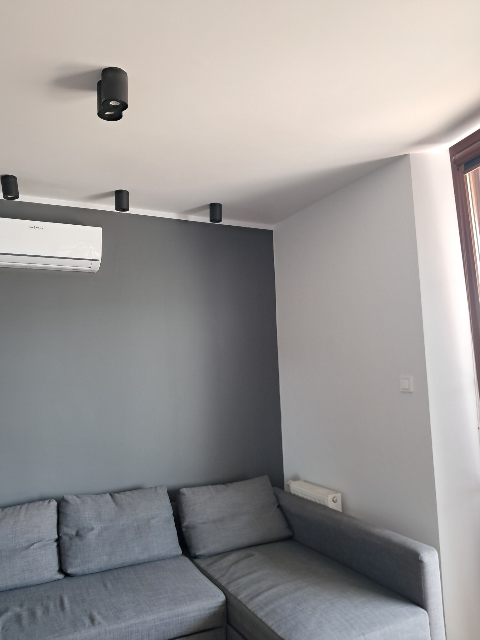 Szara ściana z klimatyzatorem Viessmann i czarnymi lampami sufitowymi. Częściowo widoczny szary narożnik i okno z brązową ramą. Minimalistyczny design wnętrza.