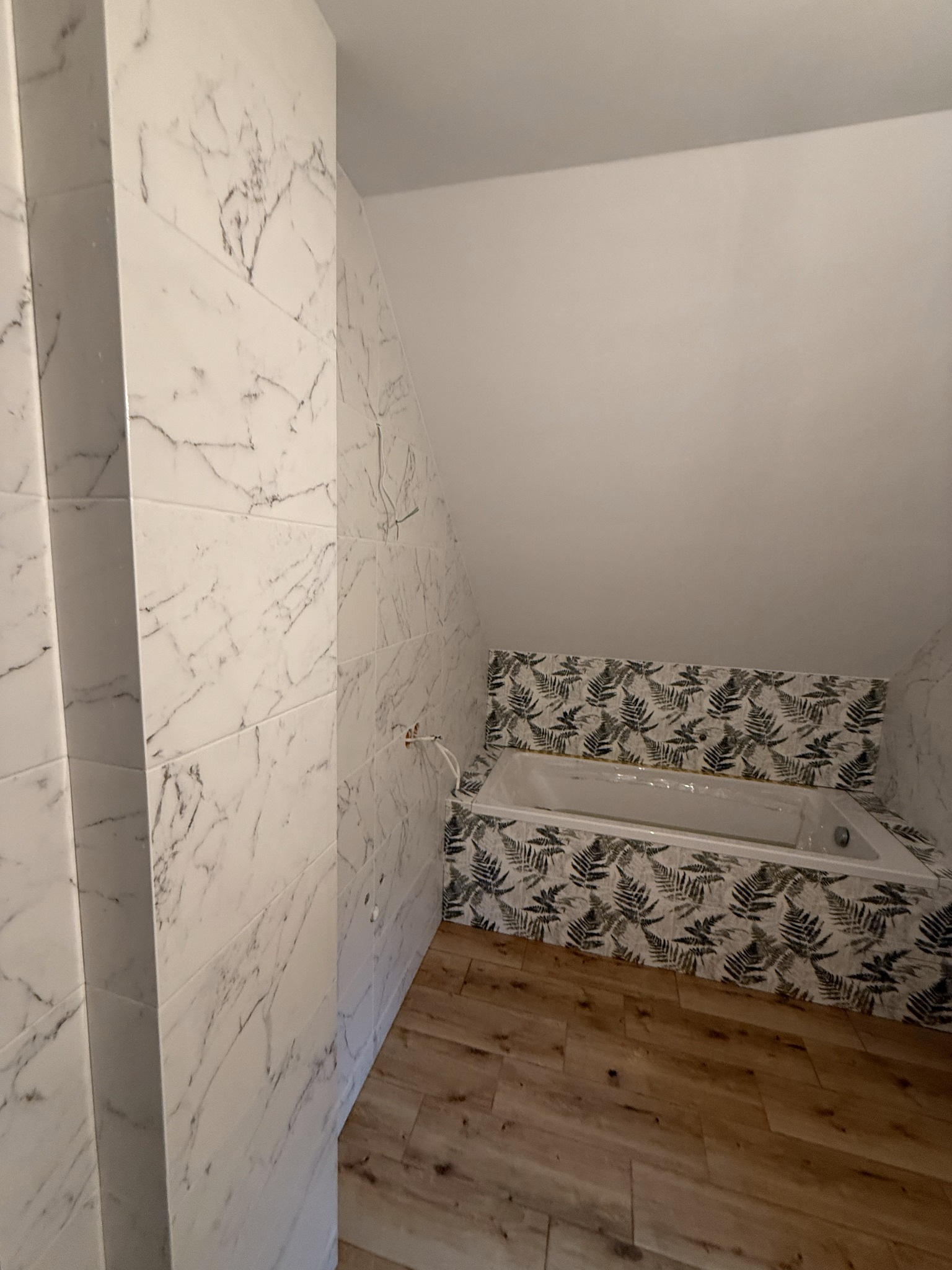 Ukończona łazienka na poddaszu: białe marmurowe płytki ścienne, wanna obudowana płytkami z motywem paproci, drewnopodobna podłoga. Widoczne wystające kable.