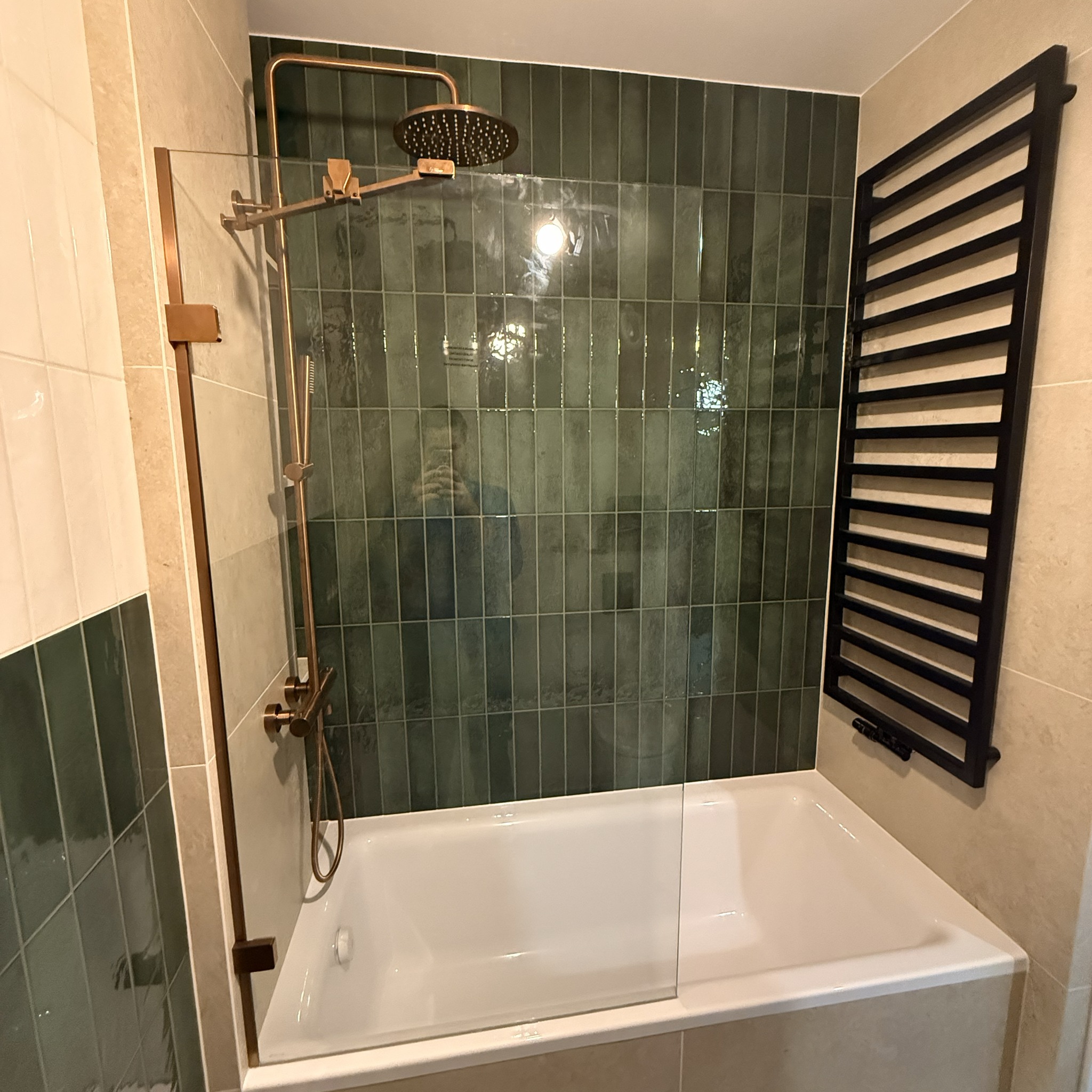 Wykończona łazienka z wanną, szklanym parawanem, deszczownicą w kolorze miedzi i czarnym grzejnikiem drabinkowym na tle zielonych, pionowych płytek.