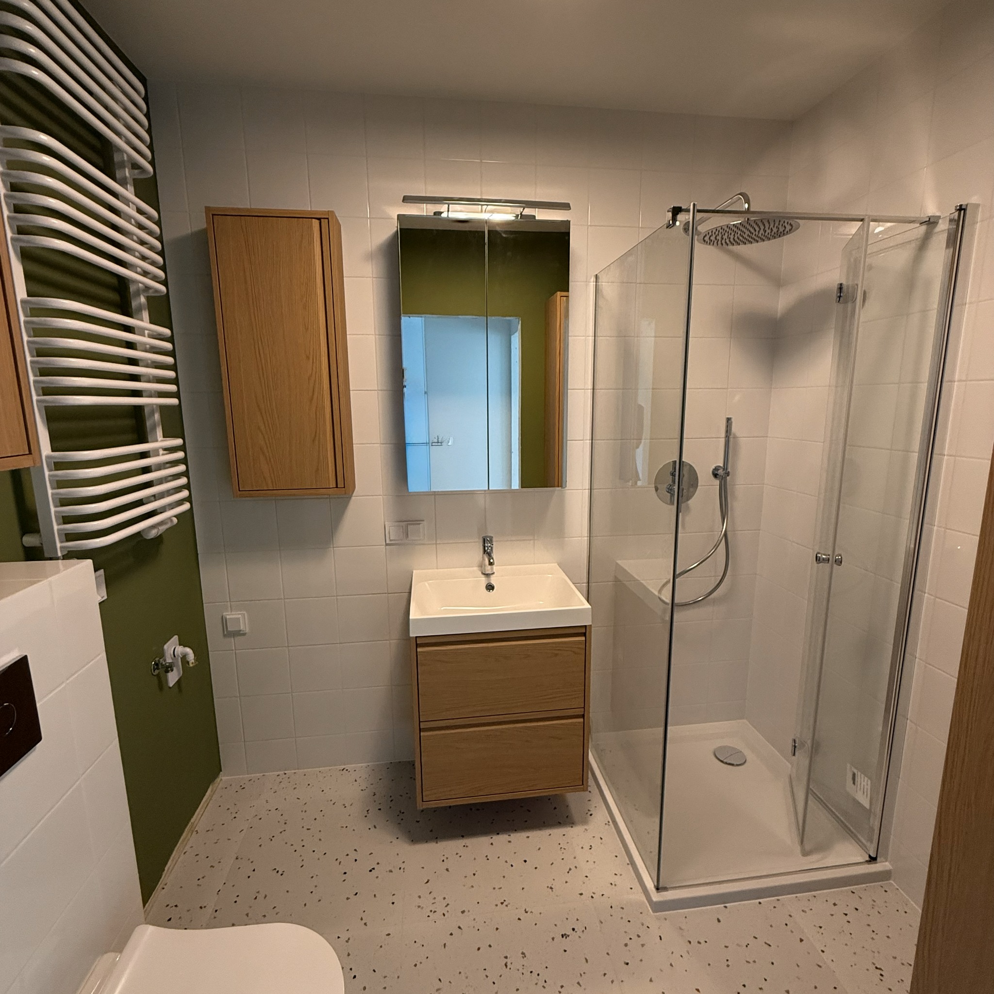 Nowoczesna łazienka z kabiną prysznicową, szafką pod umywalką z drewna i grzejnikiem drabinkowym. Białe płytki i podłoga w terazzo.