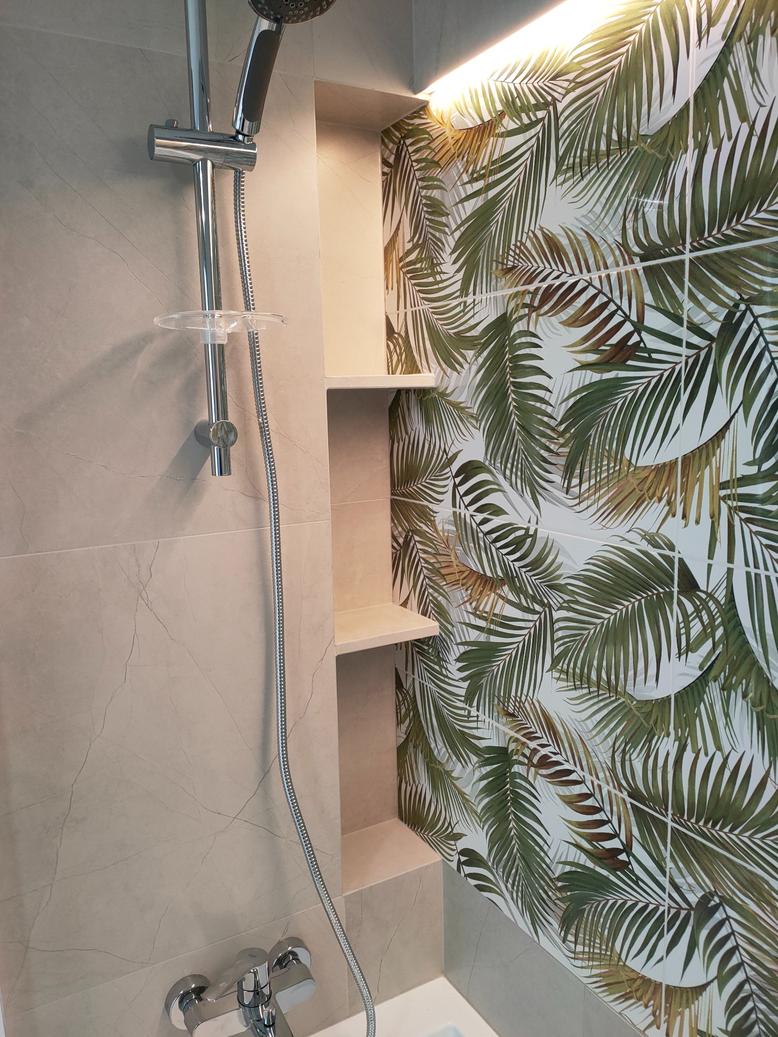 Nowoczesna kabina prysznicowa z półkami wnękowymi i płytkami w tropikalne liście. Chromowana armatura i beżowe płytki imitujące marmur tworzą spójną całość.