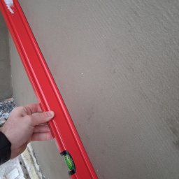 IFS SPÓŁKA Z OGRANICZONĄ ODPOWIEDZIALNOŚCIĄ - Czerwona poziomica przyłożona do szarej, świeżo nałożonej warstwy tynku na elewacji budynku. Widoczna dłoń i fragment ocieplenia. Kontrola równości powierzchni.