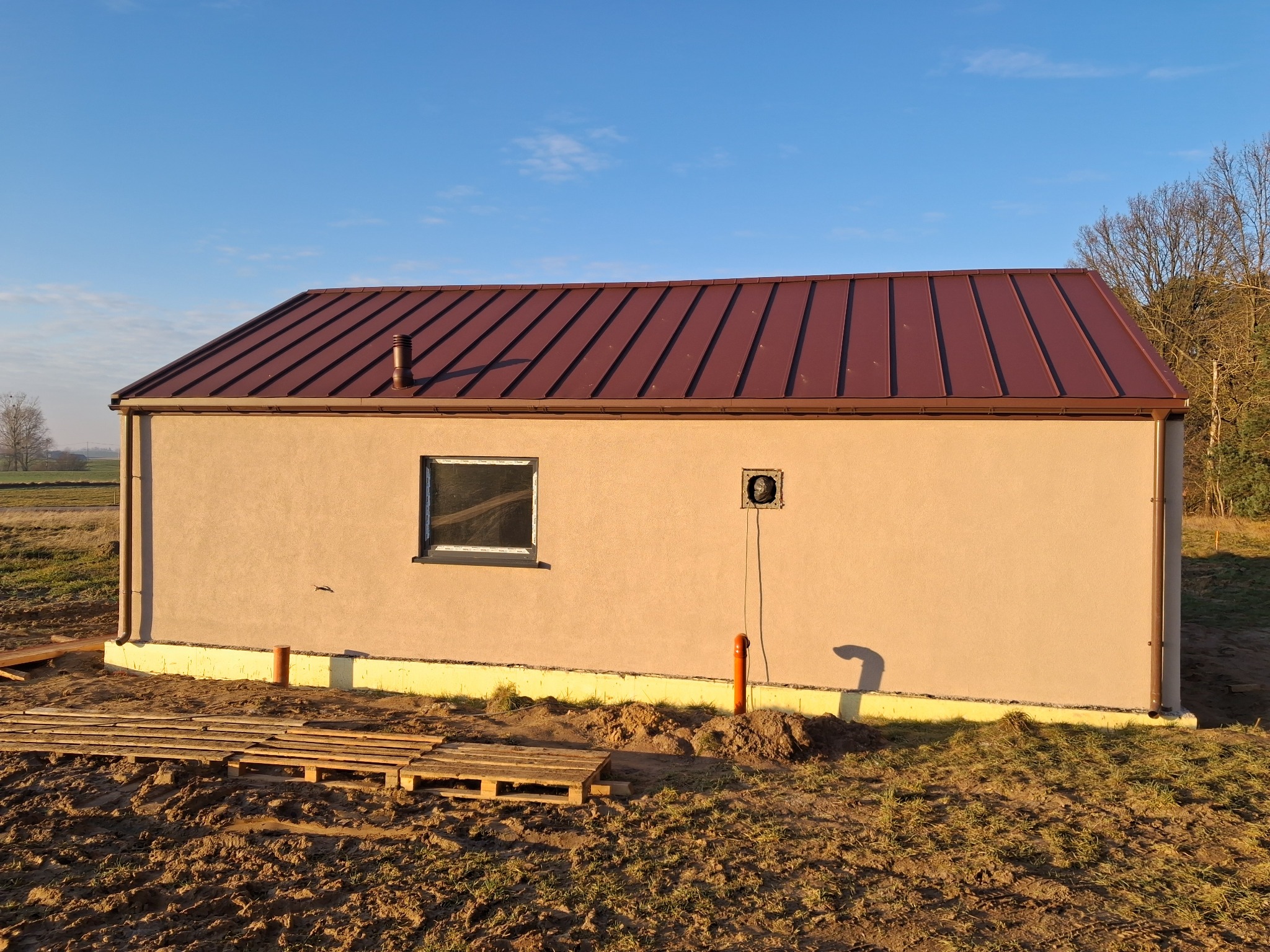 Otynkowany budynek z brązowym dachem blachodachówką, widoczne okno i rury spustowe. Żółta izolacja fundamentów. W tle drzewa i błękitne niebo.