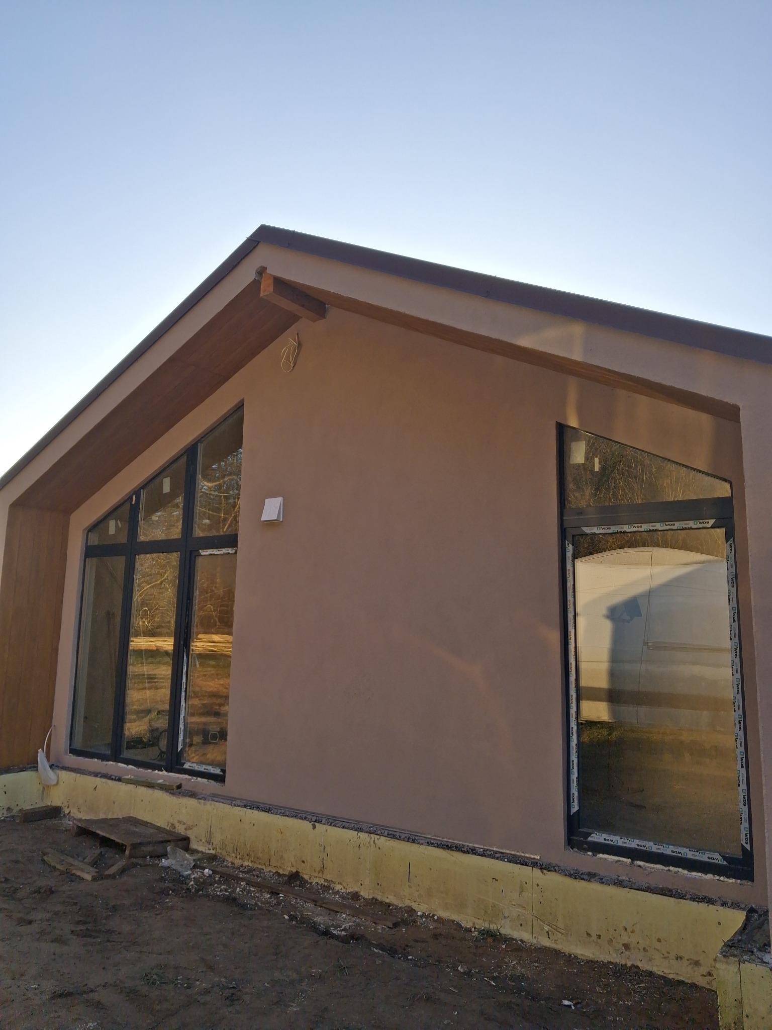 Elewacja domu jednorodzinnego z oknami o nietypowym kształcie, wykończona tynkiem w odcieniach beżu. Widoczne detale wykończeniowe i fragment terenu przed budynkiem.