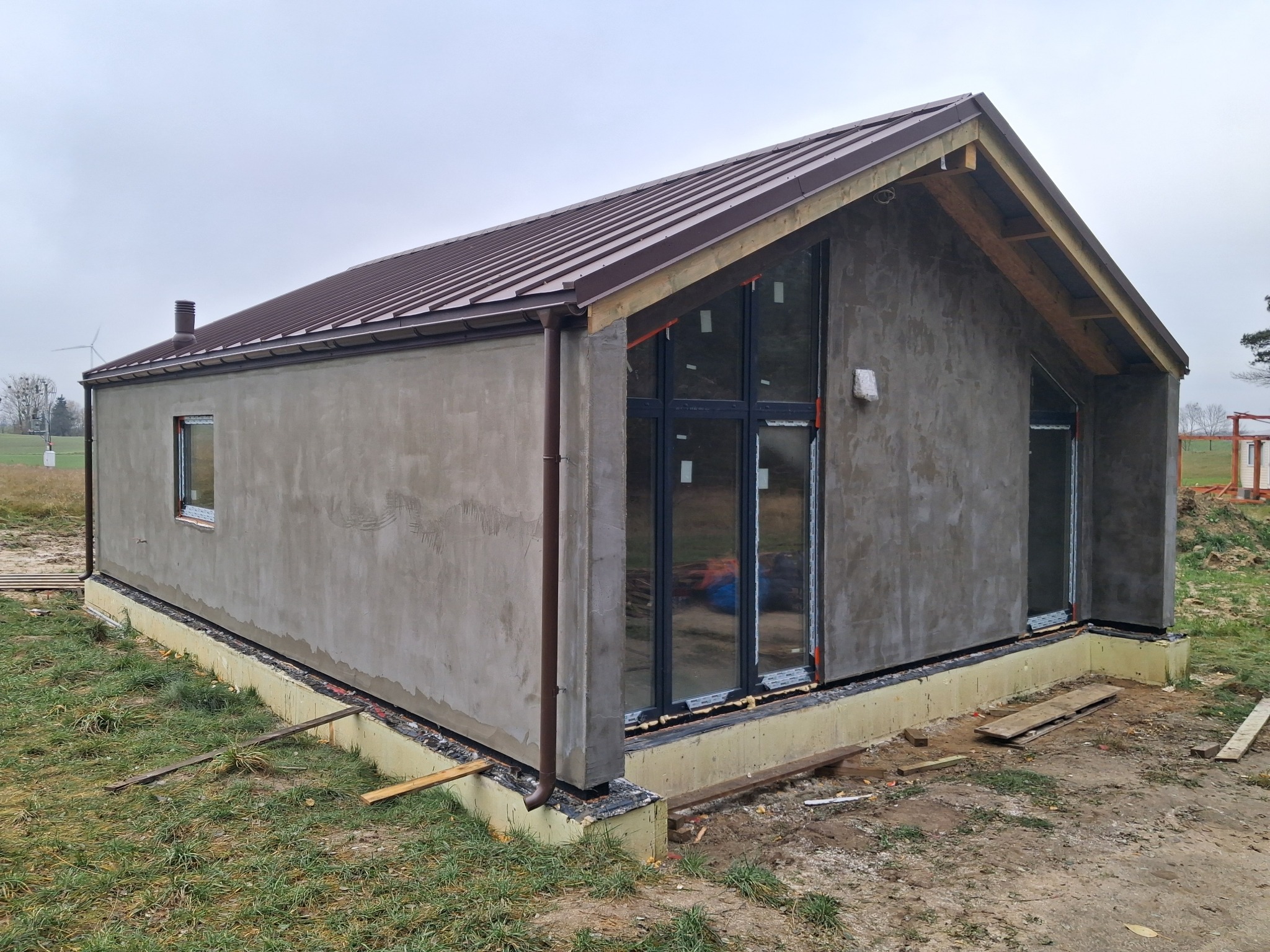 Surowy, otynkowany dom jednorodzinny z brązowym dachem z blachy, widok z zewnątrz na tle pochmurnego nieba i zielonego terenu. Widoczne okna i elementy wykończeniowe.