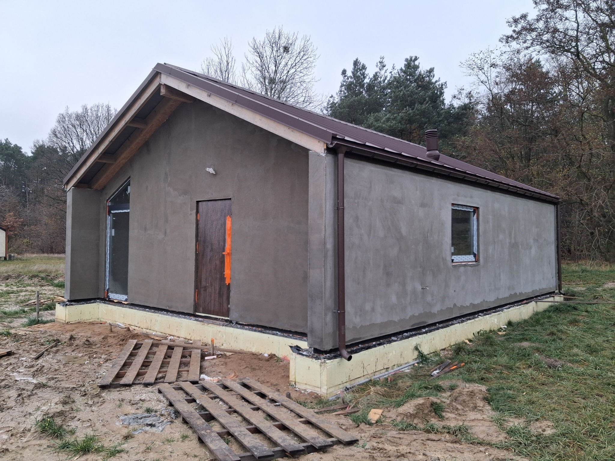 Surowy, otynkowany budynek z brązowym dachem i stolarką okienną, widoczna izolacja fundamentów, teren budowy z paletami, drzewa w tle.