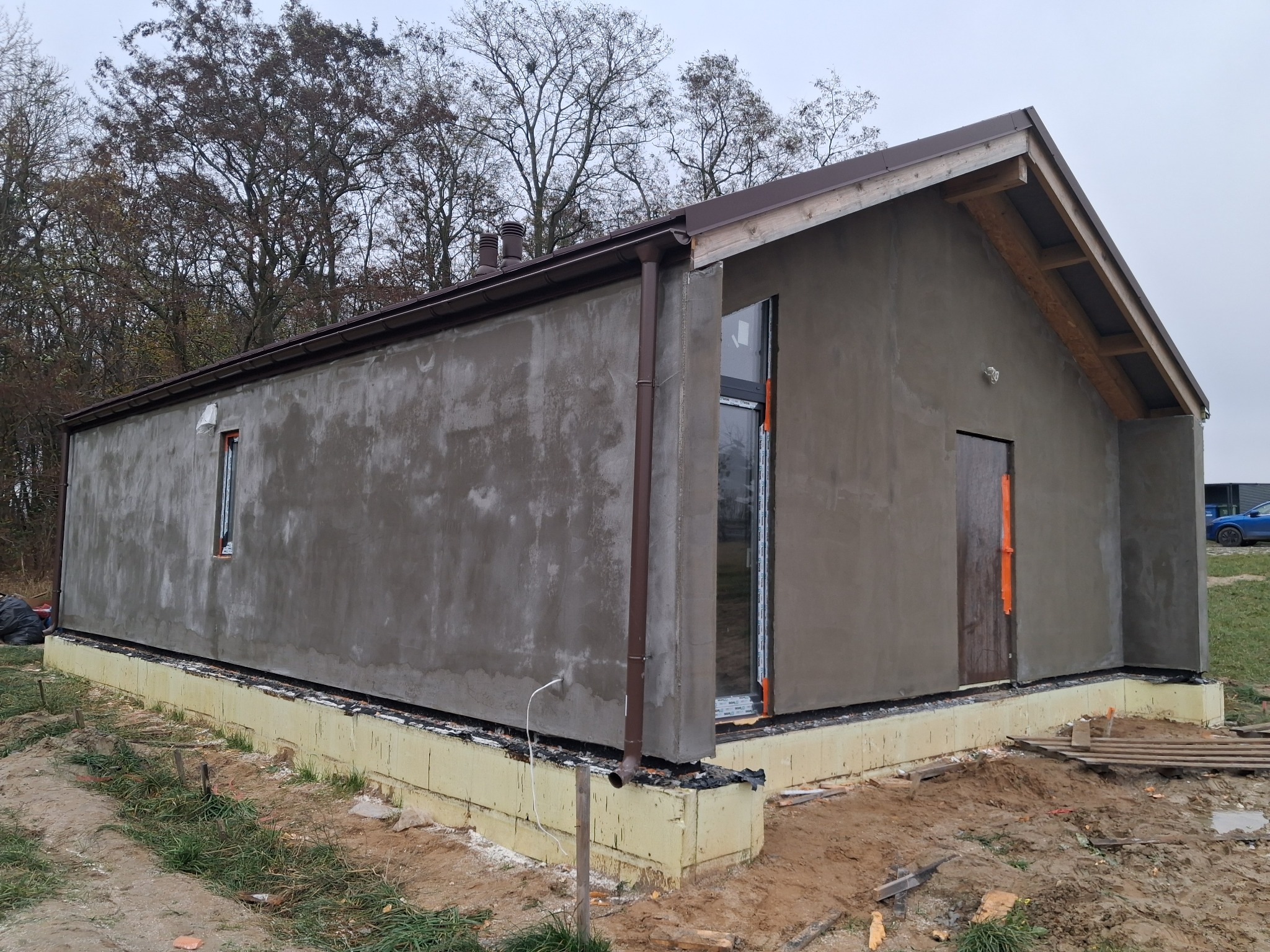 Surowy stan elewacji domu jednorodzinnego z widocznym ociepleniem fundamentów i świeżo nałożonym tynkiem. Widoczne okna i drzwi w stanie montażu.