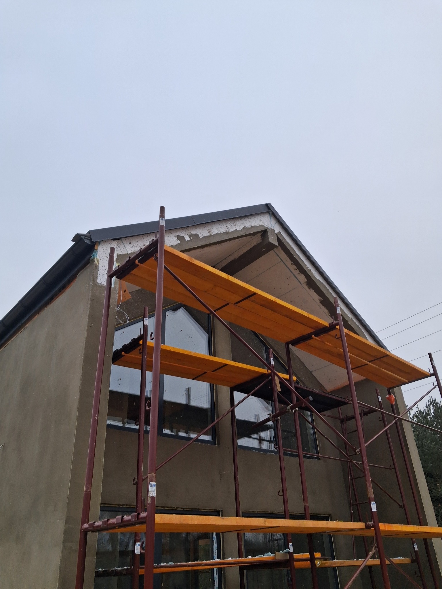 Dom w trakcie tynkowania elewacji, widoczne rusztowania i fragment ocieplenia. Szara, pochmurna pogoda.