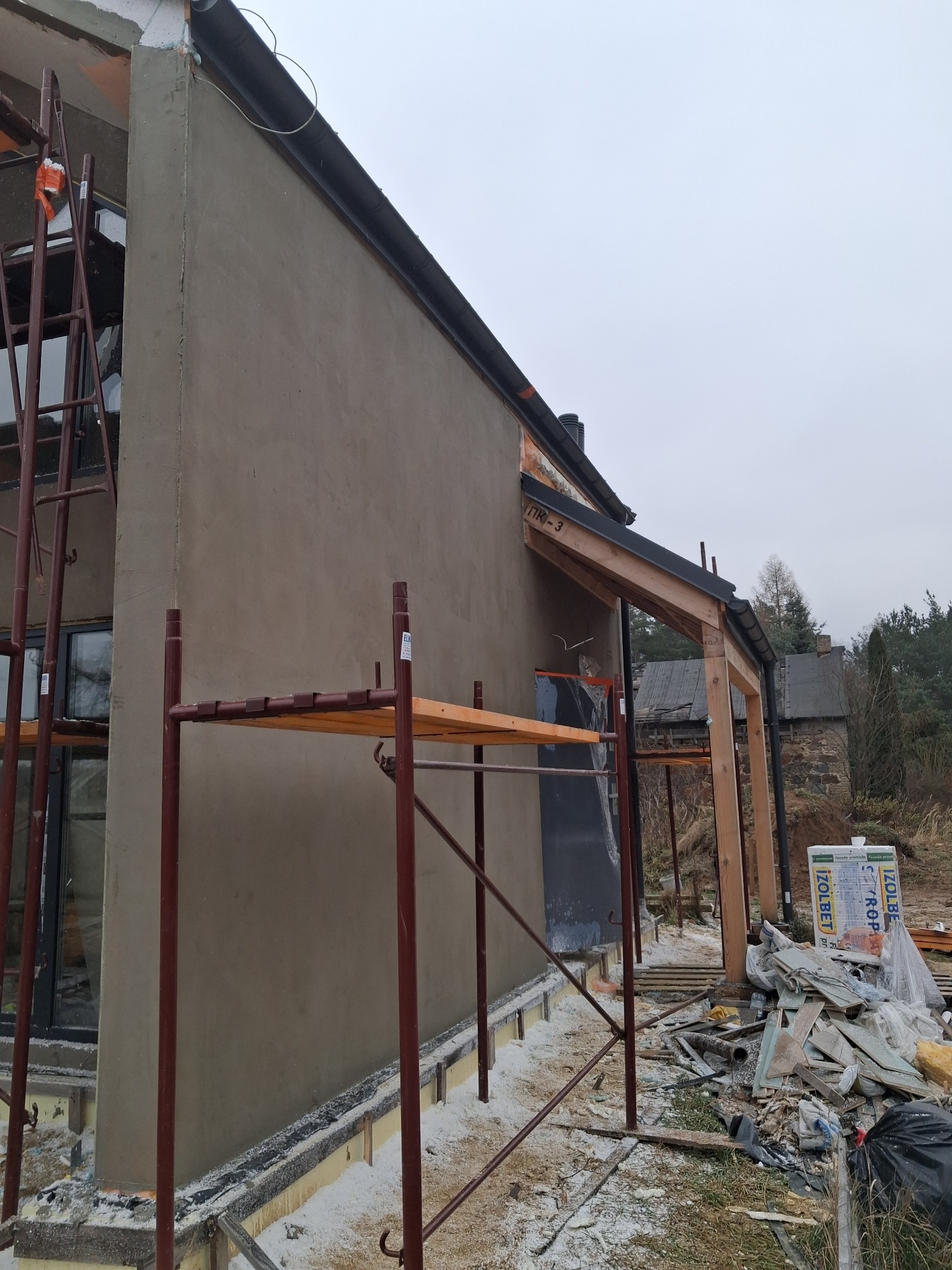 Ściana budynku w trakcie tynkowania, widoczne rusztowania i materiały budowlane. Surowy tynk w odcieniach szarości. Elementy drewniane konstrukcji dachu.