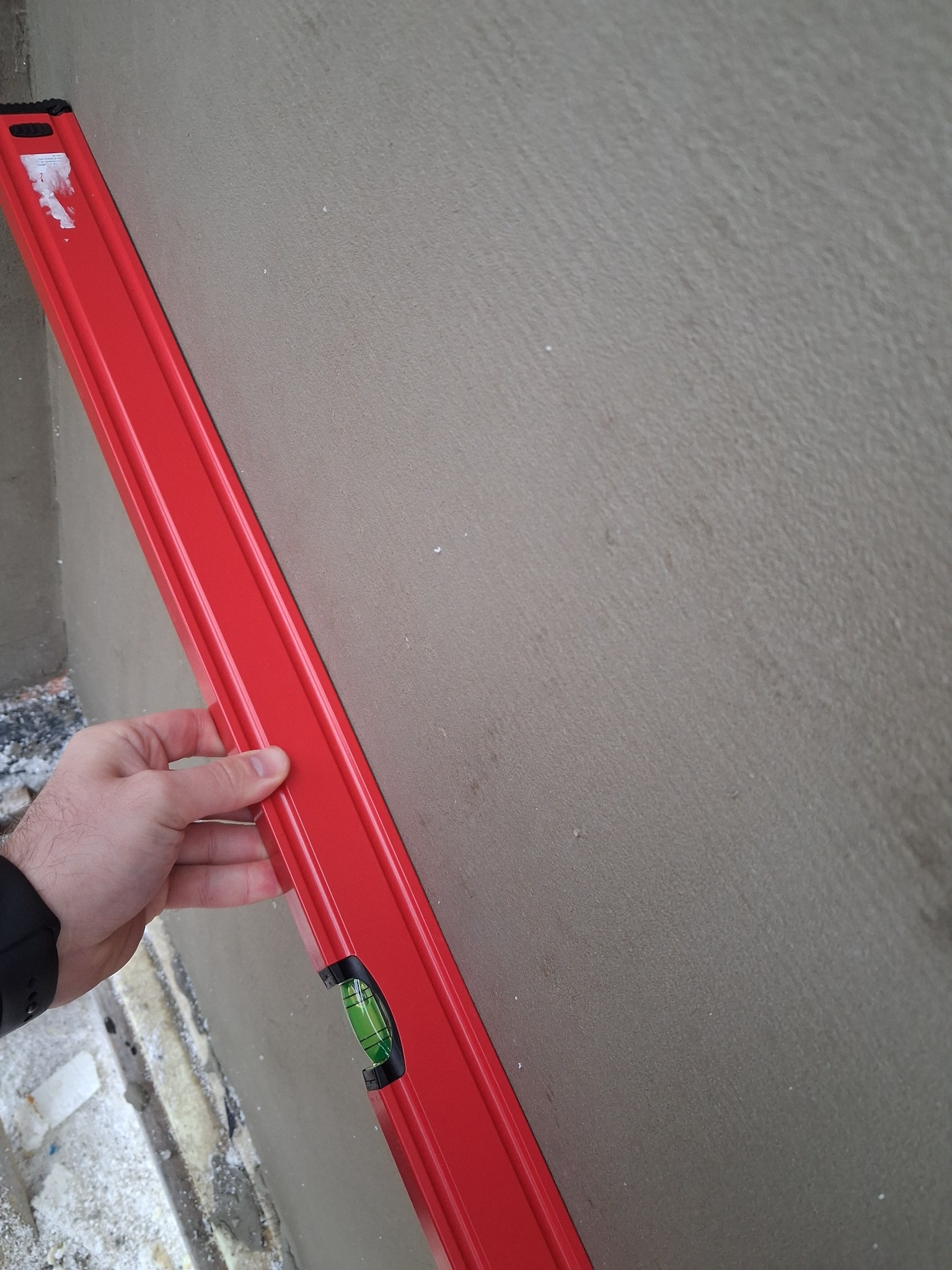 Czerwona poziomica przyłożona do szarej, świeżo nałożonej warstwy tynku na elewacji budynku. Widoczna dłoń i fragment ocieplenia. Kontrola równości powierzchni.