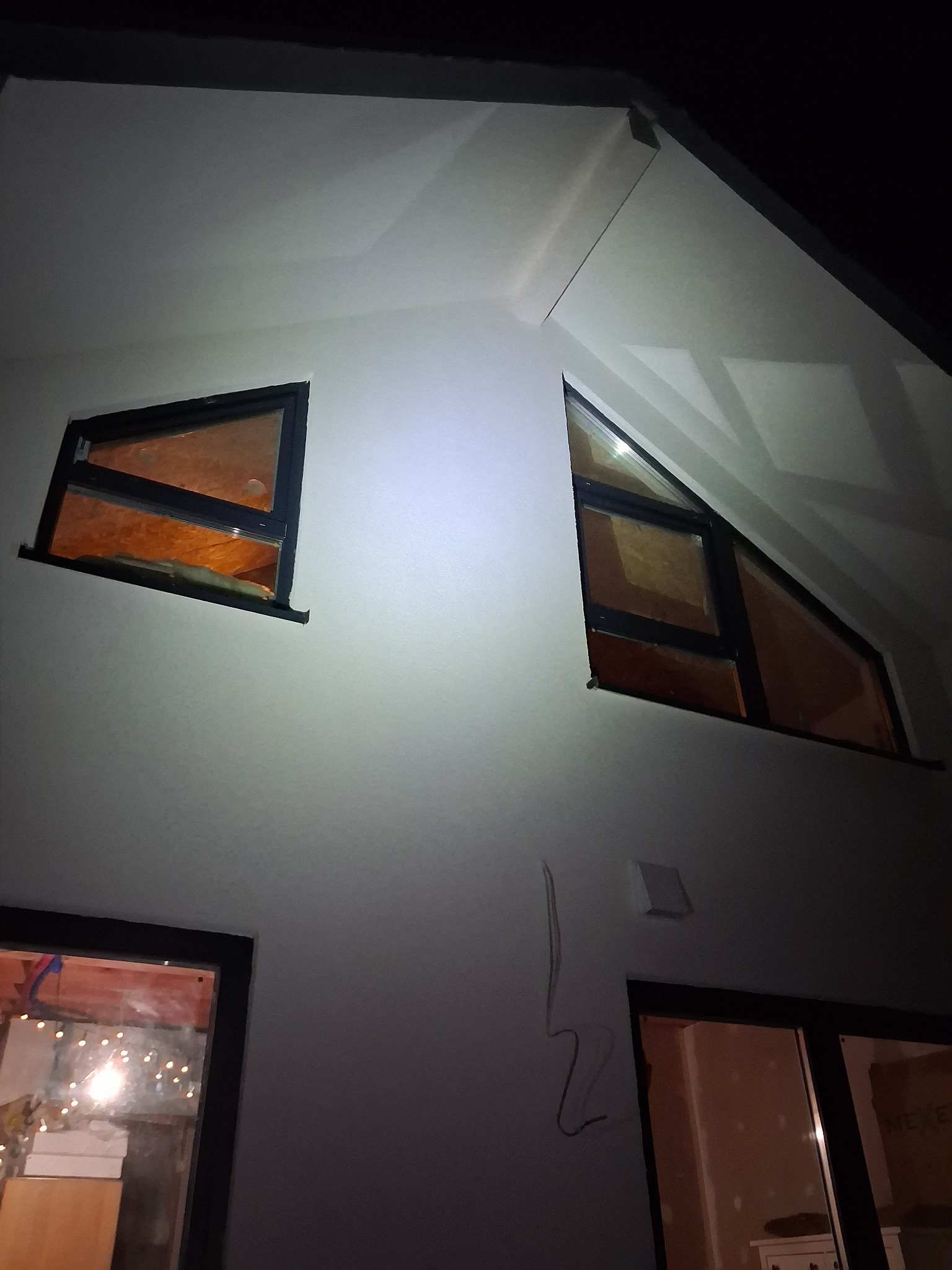 Biała elewacja domu z oknami o nietypowych kształtach (trójkątne i prostokątne) w czarnych ramach. Widoczne przewody na ścianie. Nocne ujęcie.