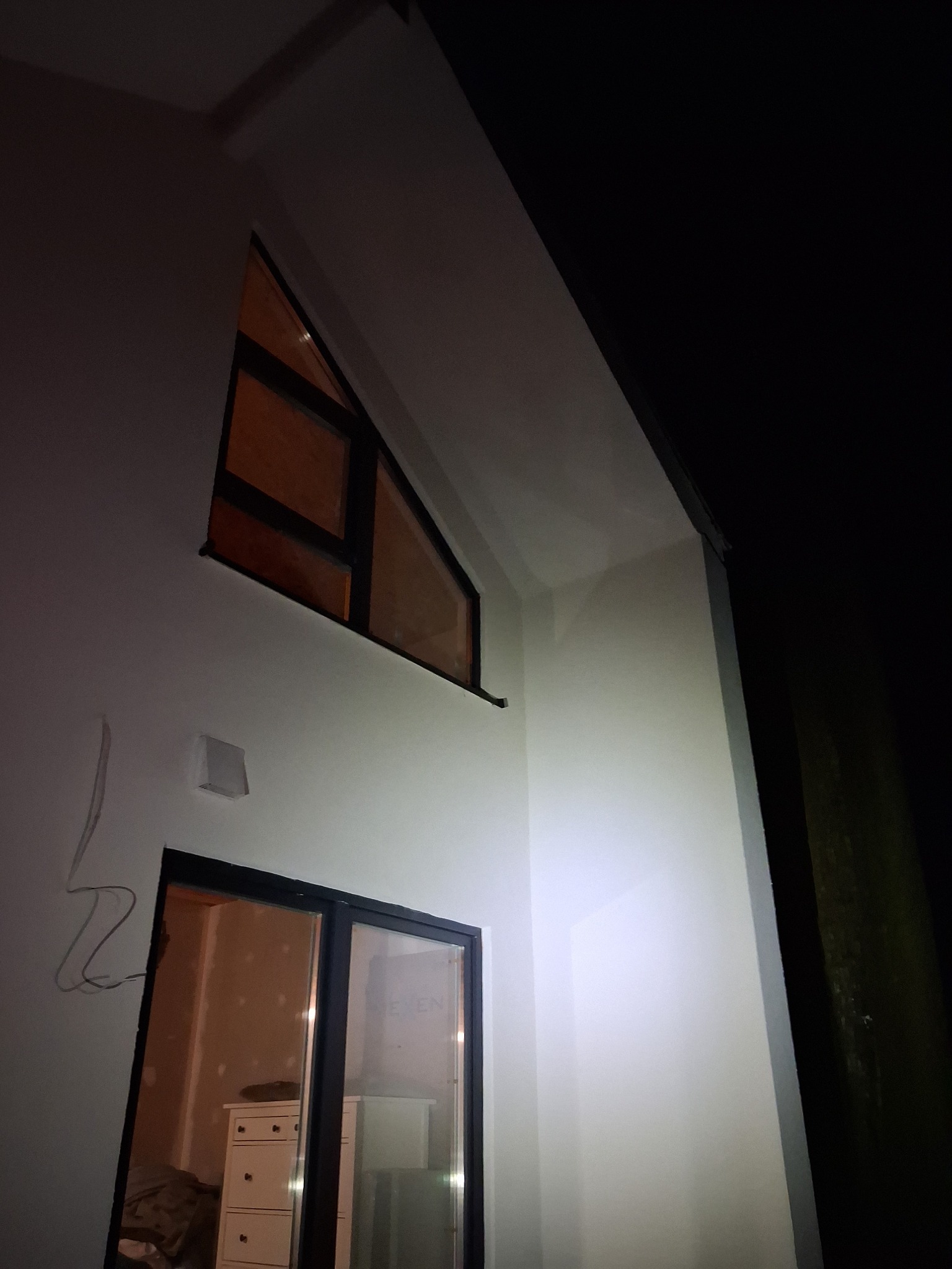 Biała elewacja domu z oknem trójkątnym i przesuwnymi drzwiami balkonowymi, widok z dołu, nocne oświetlenie, widoczne przewody elektryczne.