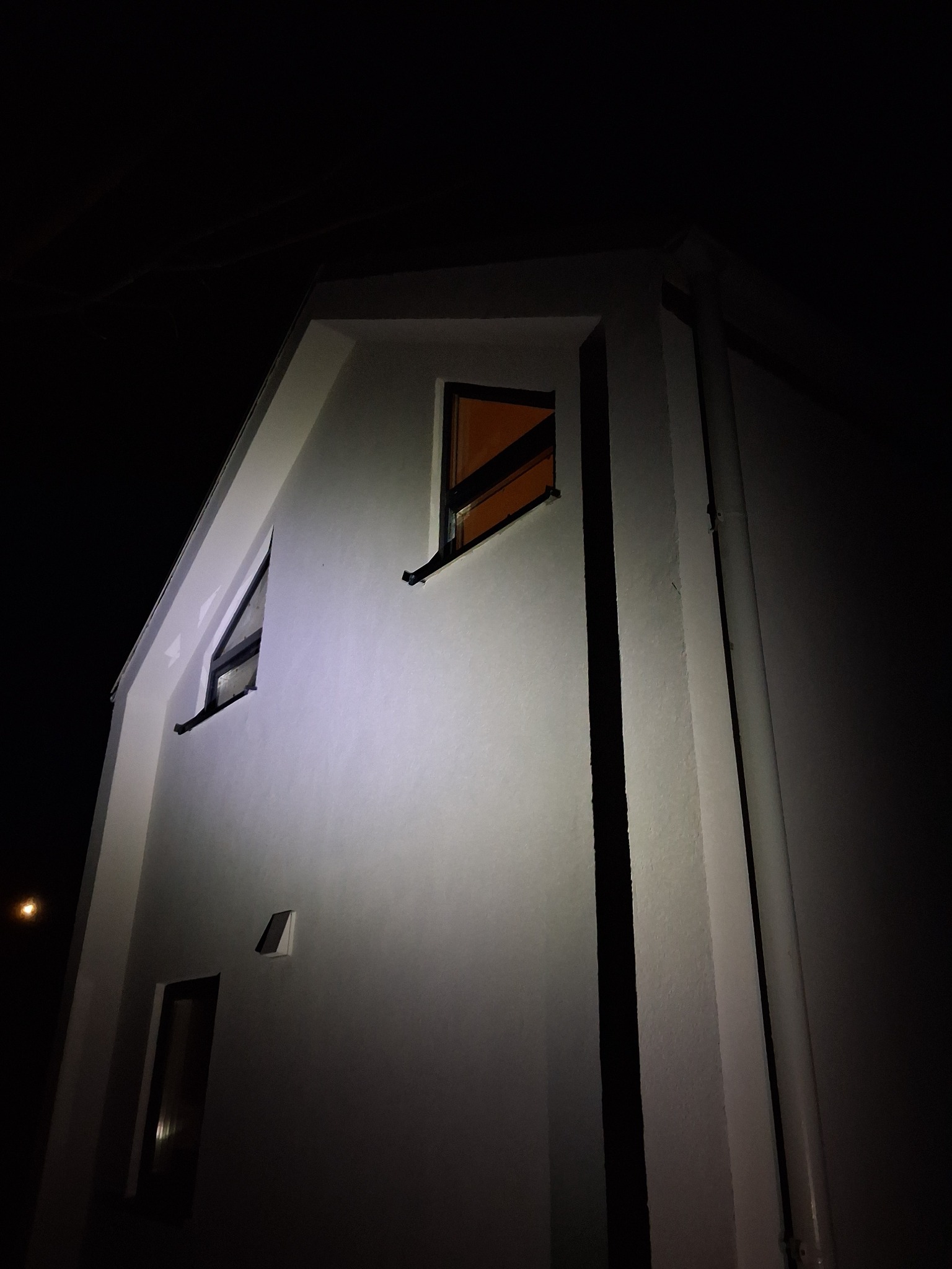 Nocne ujęcie fragmentu białej elewacji domu z oknami, oświetlonej z dołu, z widocznymi rurami spustowymi. Minimalistyczny design budynku.