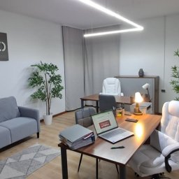 JAGIMO Sp. z o.o. - Nowoczesne biuro z drewnianym znakiem 'JAGIMO', szarą sofą, biurkami i roślinami doniczkowymi. Laptop na biurku z widoczną stroną internetową. Jasne, przestronne wnętrze.