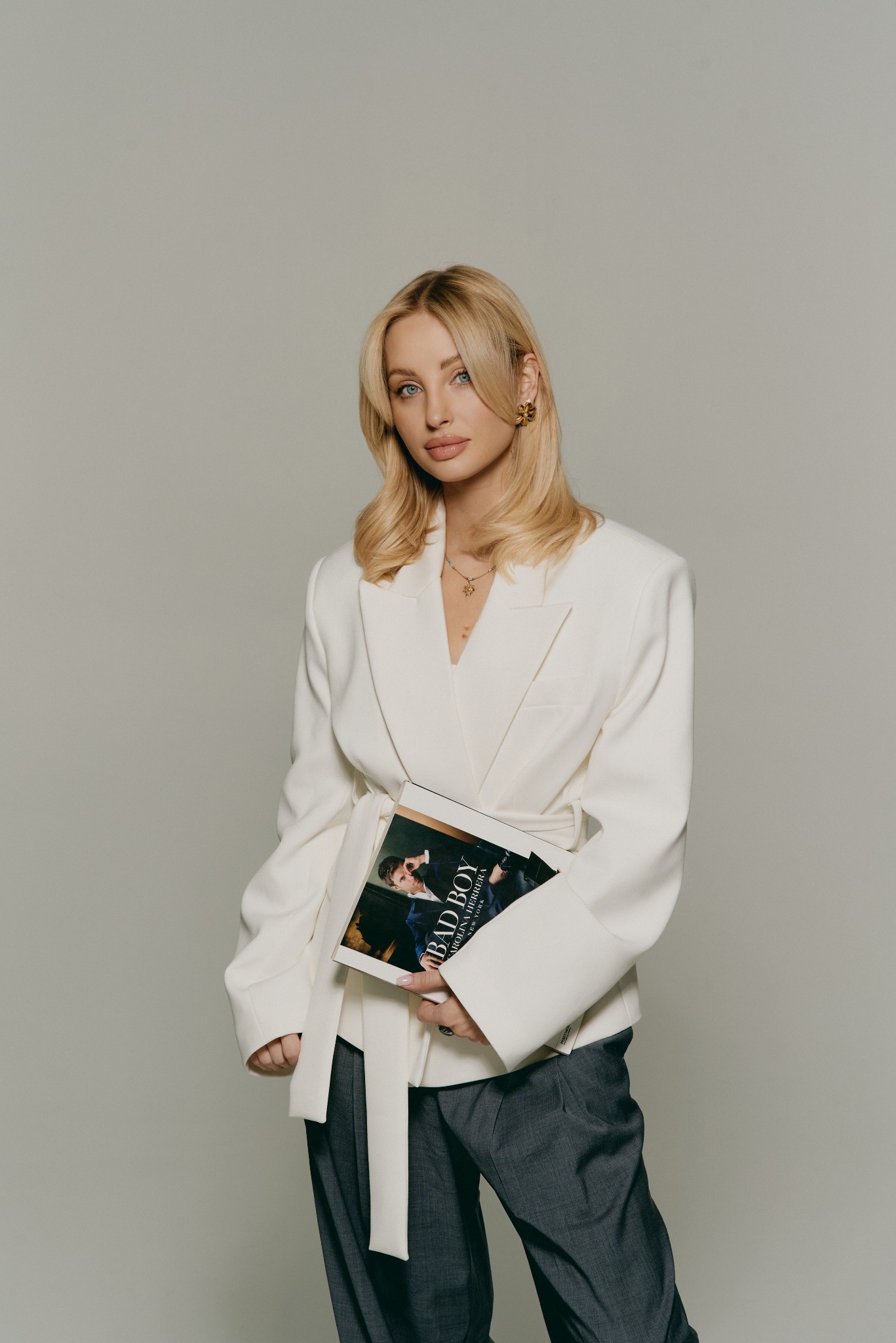 Blondwłosa kobieta w białym żakiecie trzymająca magazyn modowy 'BAD BOY' na neutralnym tle. Subtelny makijaż, złota biżuteria i luźne spodnie dopełniają stylizację.