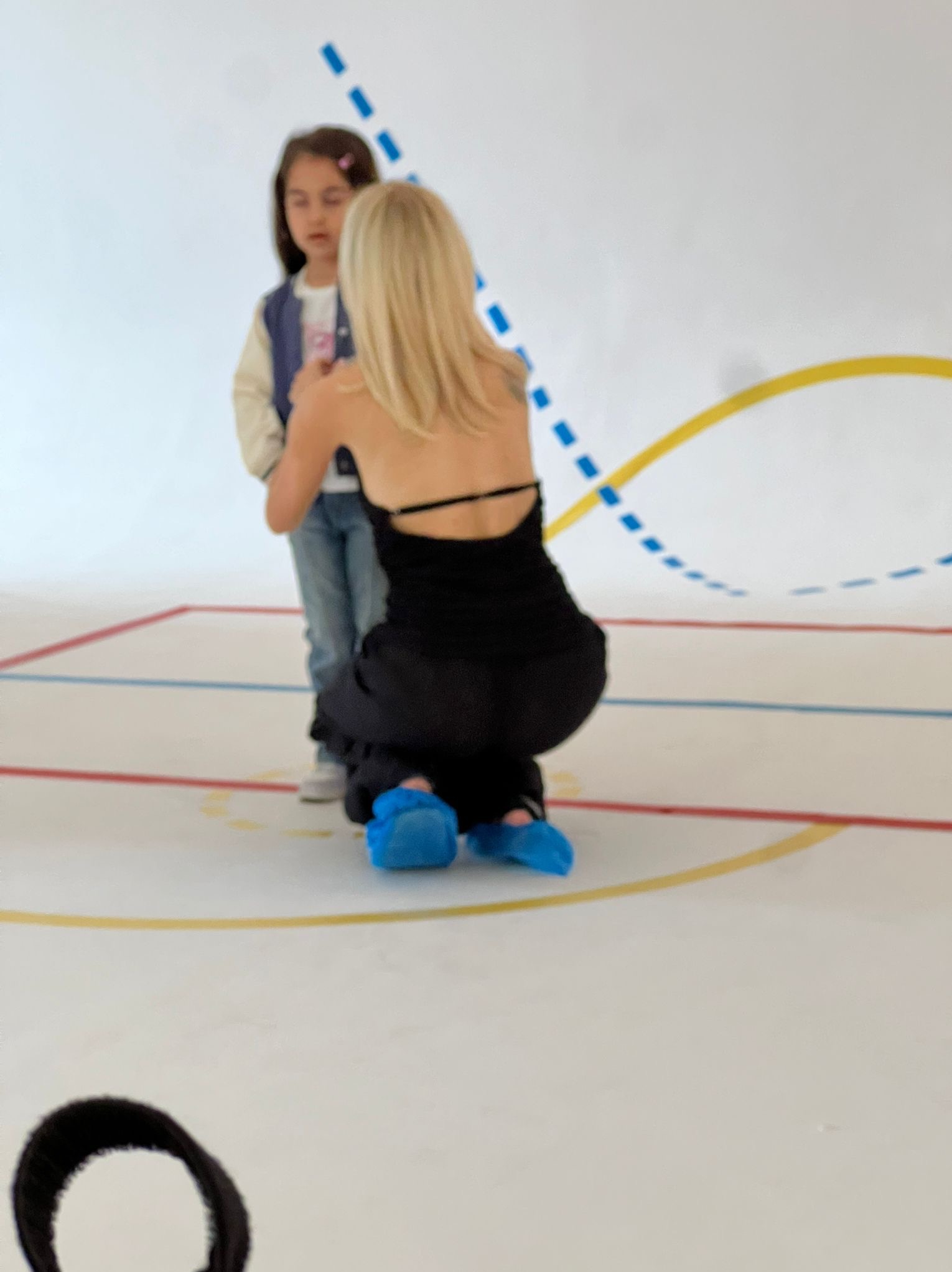 Kobieta w czarnym kombinezonie, w pozycji kucznej, poprawia ubranie dziewczynce na tle białego studia z kolorowymi liniami. Przygotowania do sesji zdjęciowej.