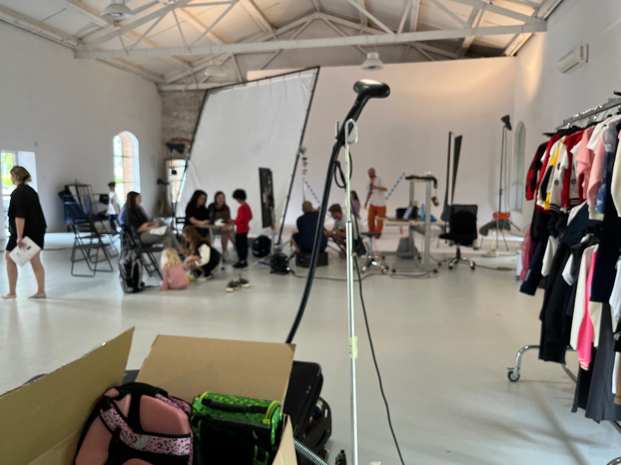 Sesja zdjęciowa w studio: grupa osób, ubrania na wieszakach, oświetlenie studyjne, kartonowe pudło na pierwszym planie. Przygotowania do sesji.