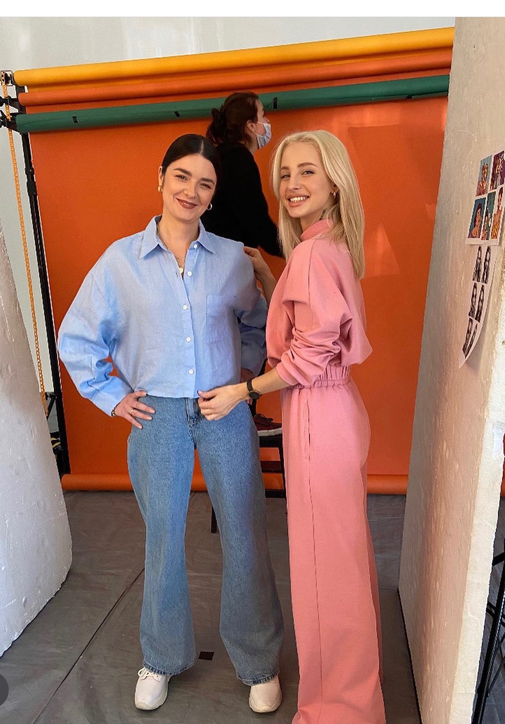 Dwie uśmiechnięte kobiety pozują na tle pomarańczowego tła w studio. Jedna w niebieskiej koszuli i dżinsach, druga w różowym dresie. Stylistka poprawia ubranie.