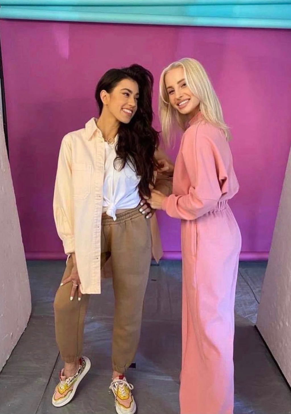 Dwie uśmiechnięte kobiety w modnych, pastelowych stylizacjach na tle fioletowego studia. Brunetka w beżach, blondynka w różu. Sesja zdjęciowa.