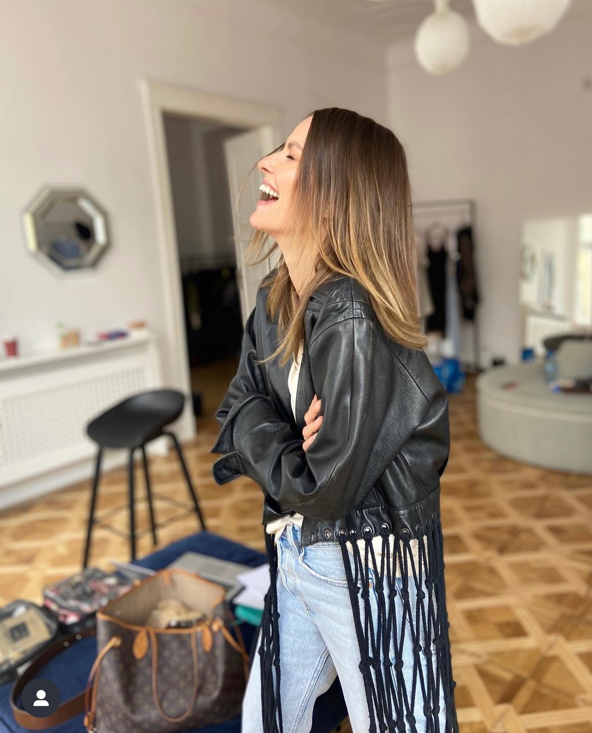 Uśmiechnięta blondynka w skórzanej kurtce z frędzlami i jasnych jeansach, stoi w jasnym wnętrzu z torbą i wieszakiem na ubrania w tle. Sesja stylizacyjna.