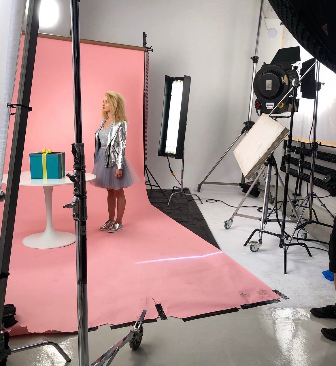 Sesja zdjęciowa: blond modelka w srebrnej kurtce i tiulowej sukience pozuje na różowym tle obok prezentu, otoczona sprzętem oświetleniowym w studiu.