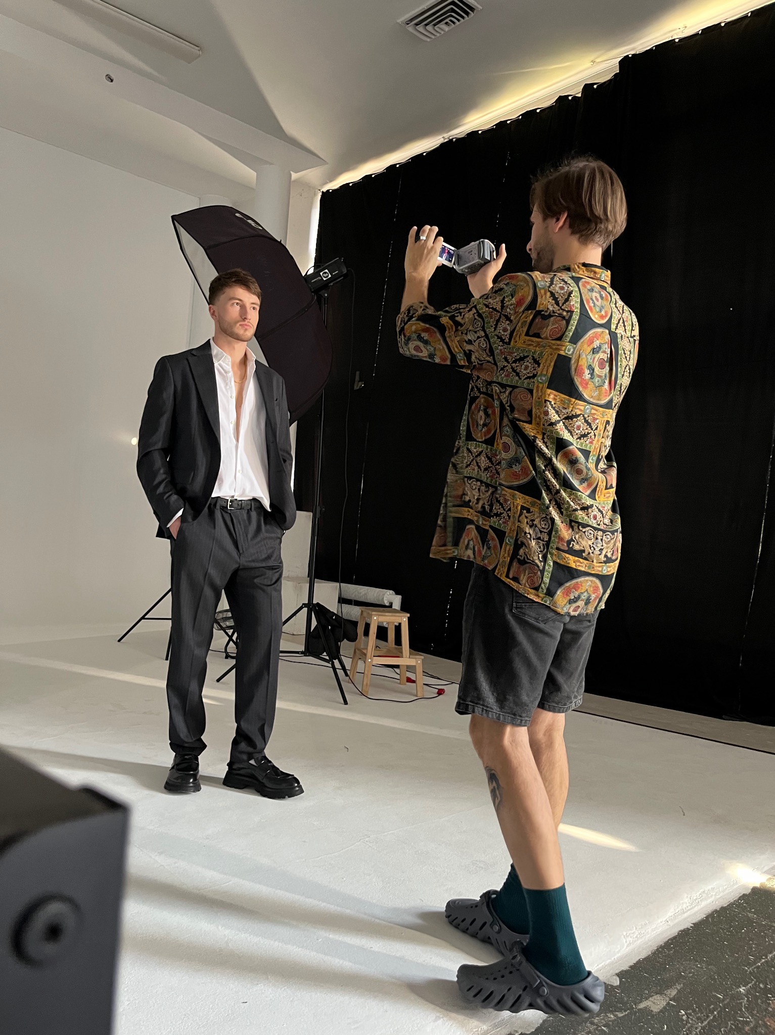 Sesja zdjęciowa: Mężczyzna w garniturze pozuje w studiu, fotograf rejestruje ujęcie kamerą. Widoczne oświetlenie studyjne i tło. Uwagę zwraca stylizacja modela.