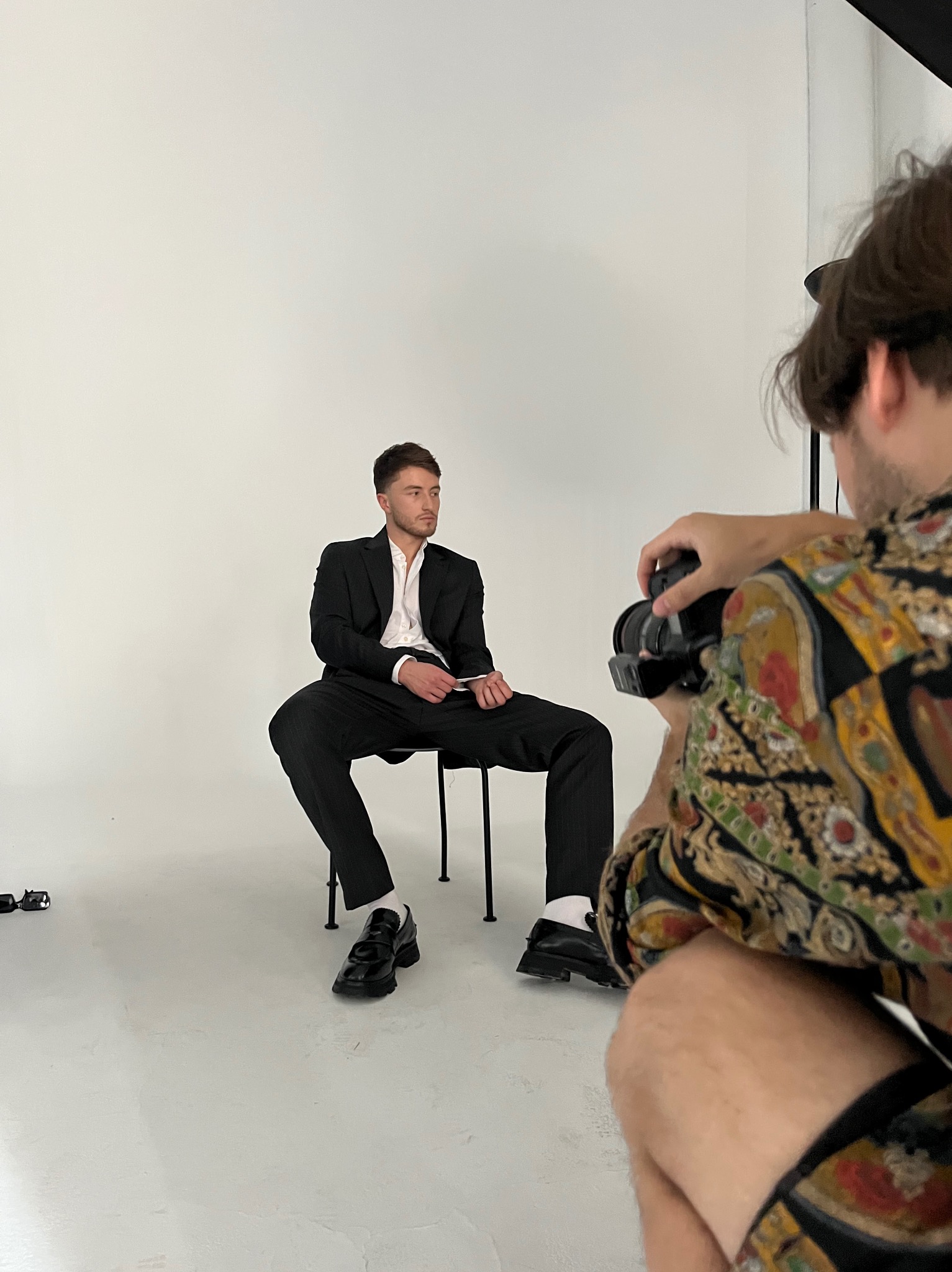 Sesja zdjęciowa: Mężczyzna w czarnym garniturze pozuje siedząc na krześle, fotograf uchwycony w kadrze z aparatem, białe tło, studyjne oświetlenie.