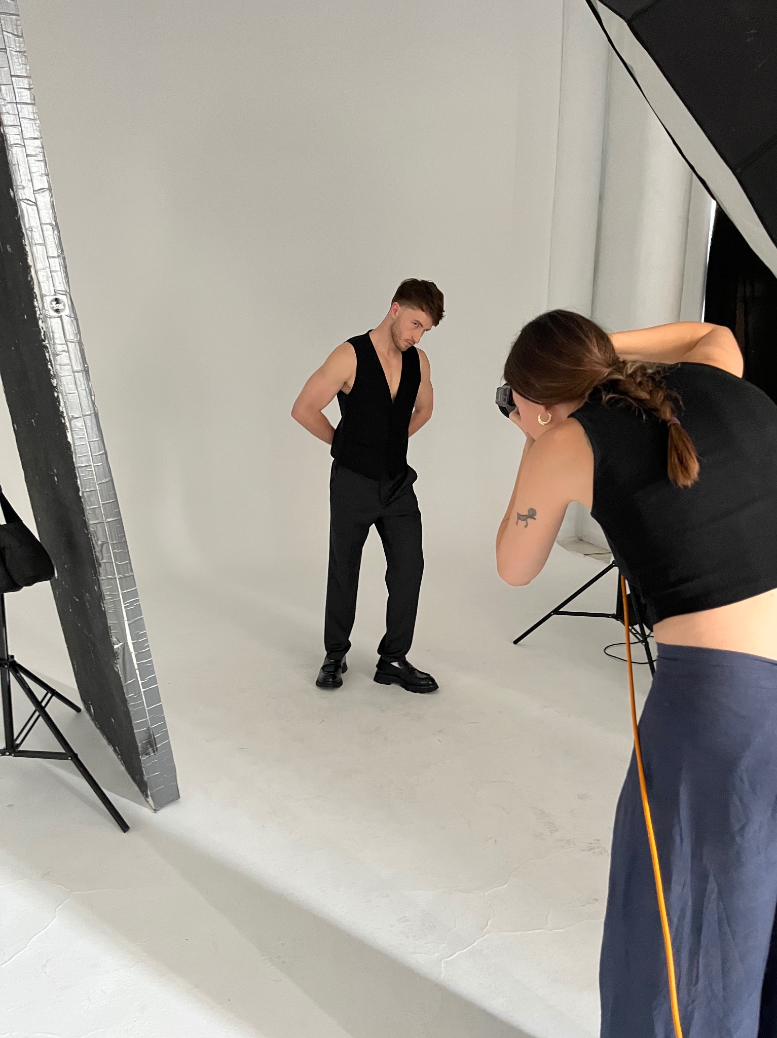 Sesja zdjęciowa: mężczyzna w czarnym stroju pozuje, fotografka robi zdjęcia w studio z białym tłem i oświetleniem studyjnym. Widoczny softbox i blendy.