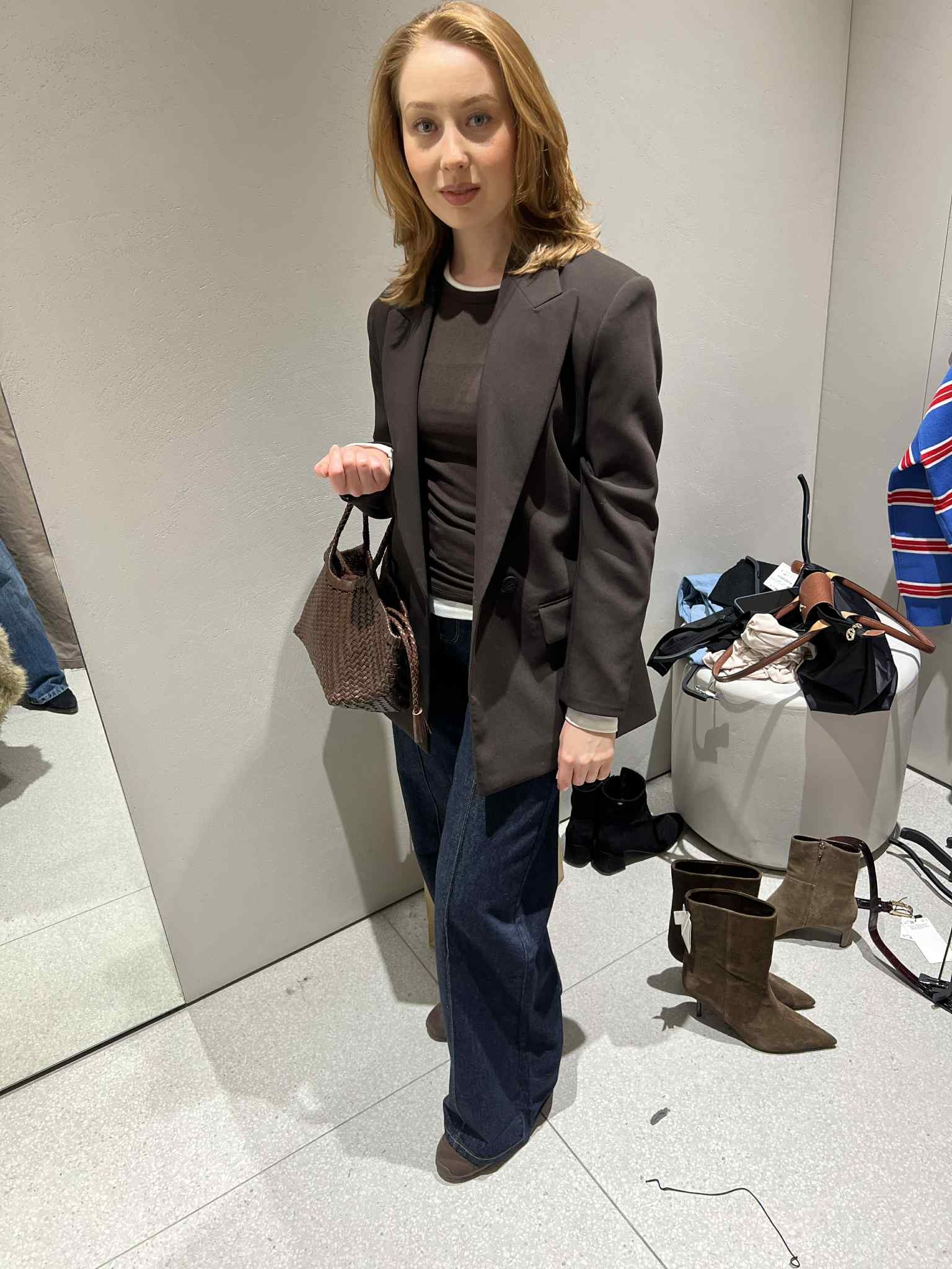 Kobieta w przymierzalni prezentuje stylizację: brązowy żakiet, ciemne dżinsy i torebka z plecionki. W tle lustro i akcesoria.