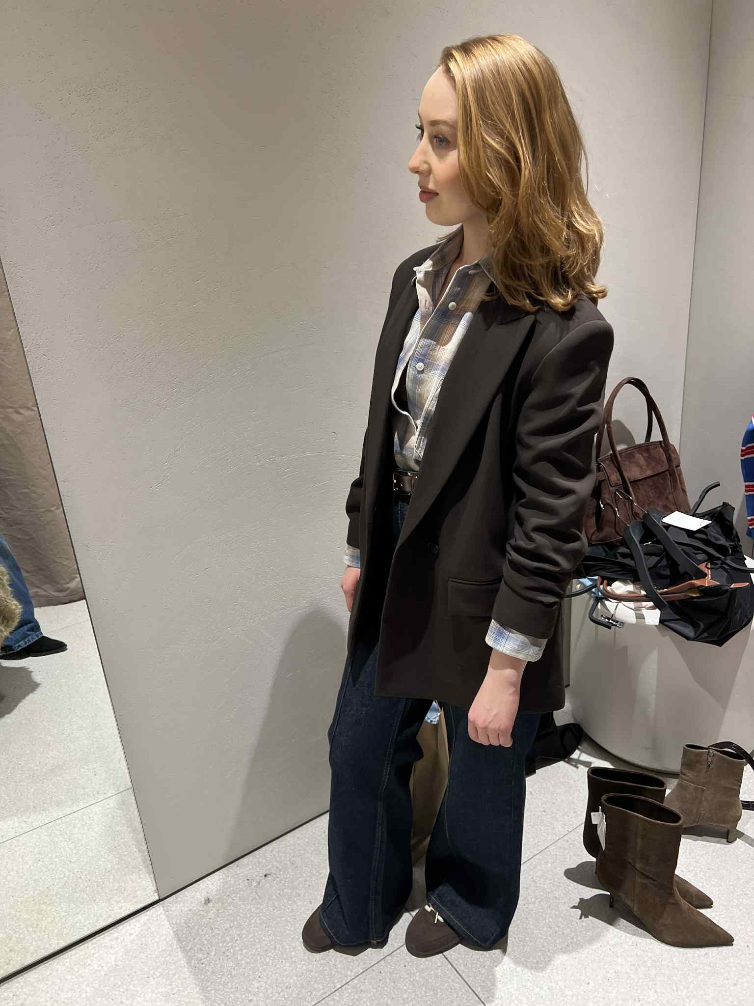 Kobieta w brązowej marynarce i dżinsach typu bootcut, pozująca w przymierzalni. Widoczne brązowe botki i torba. Stonowana kolorystyka, naturalne światło.