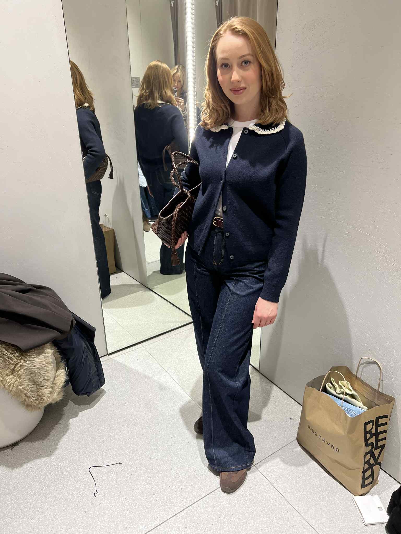 Kobieta w przymierzalni prezentuje stylizację: ciemne jeansy, kardigan z ozdobnym kołnierzykiem i torebkę. W tle lustro i torba z zakupami.