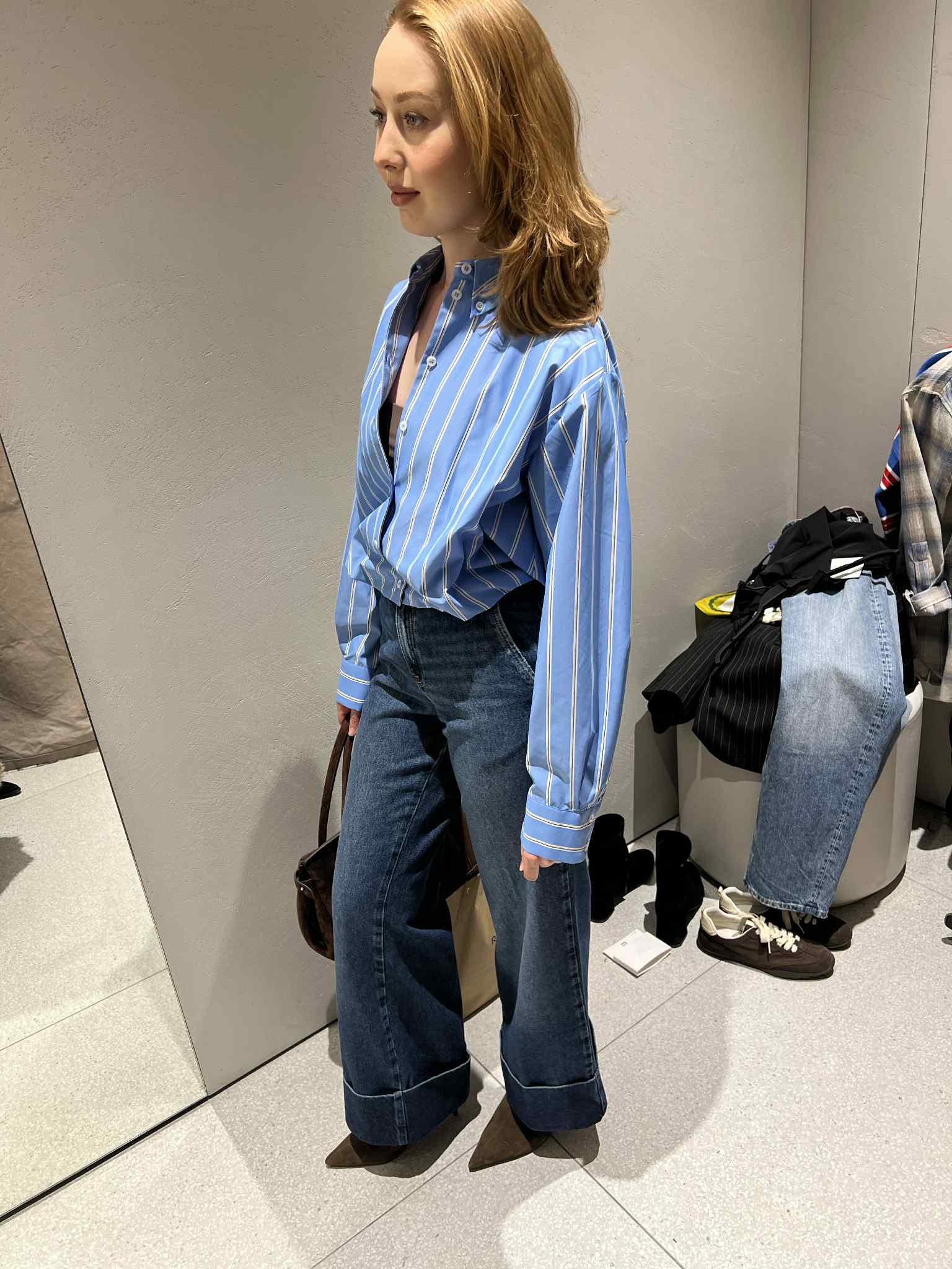 Kobieta w przymierzalni prezentuje stylizację: luźna, błękitna koszula w paski i szerokie jeansy z podwiniętymi nogawkami. W tle ubrania i lustro.