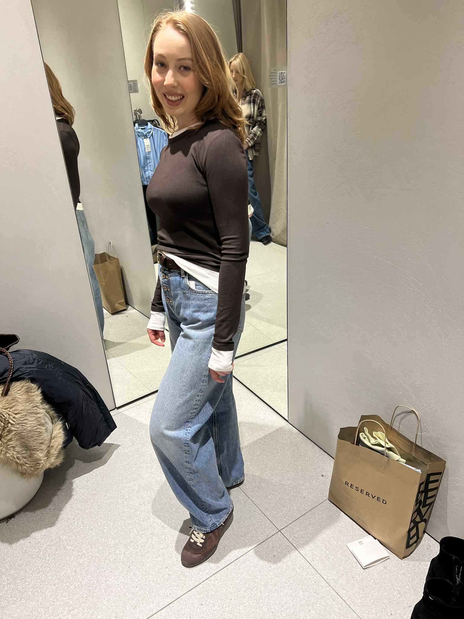 Uśmiechnięta kobieta w przymierzalni, prezentująca szerokie jeansy z wysokim stanem i brązową bluzkę. Widoczna torba Reserved i lustro.