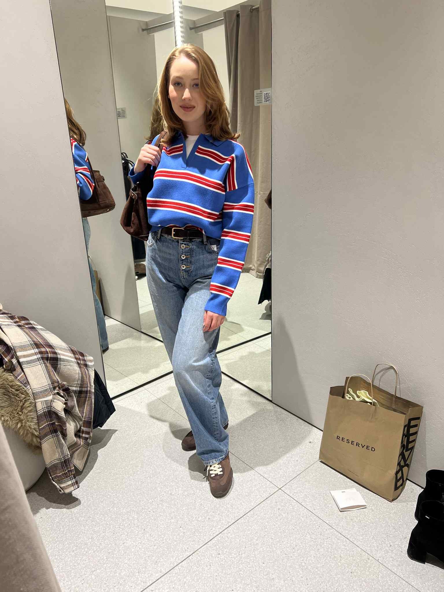Młoda kobieta w przymierzalni, prezentuje stylizację: niebieski sweter w paski, jeansy z wysokim stanem i brązowe buty. W tle lustro i torba Reserved.