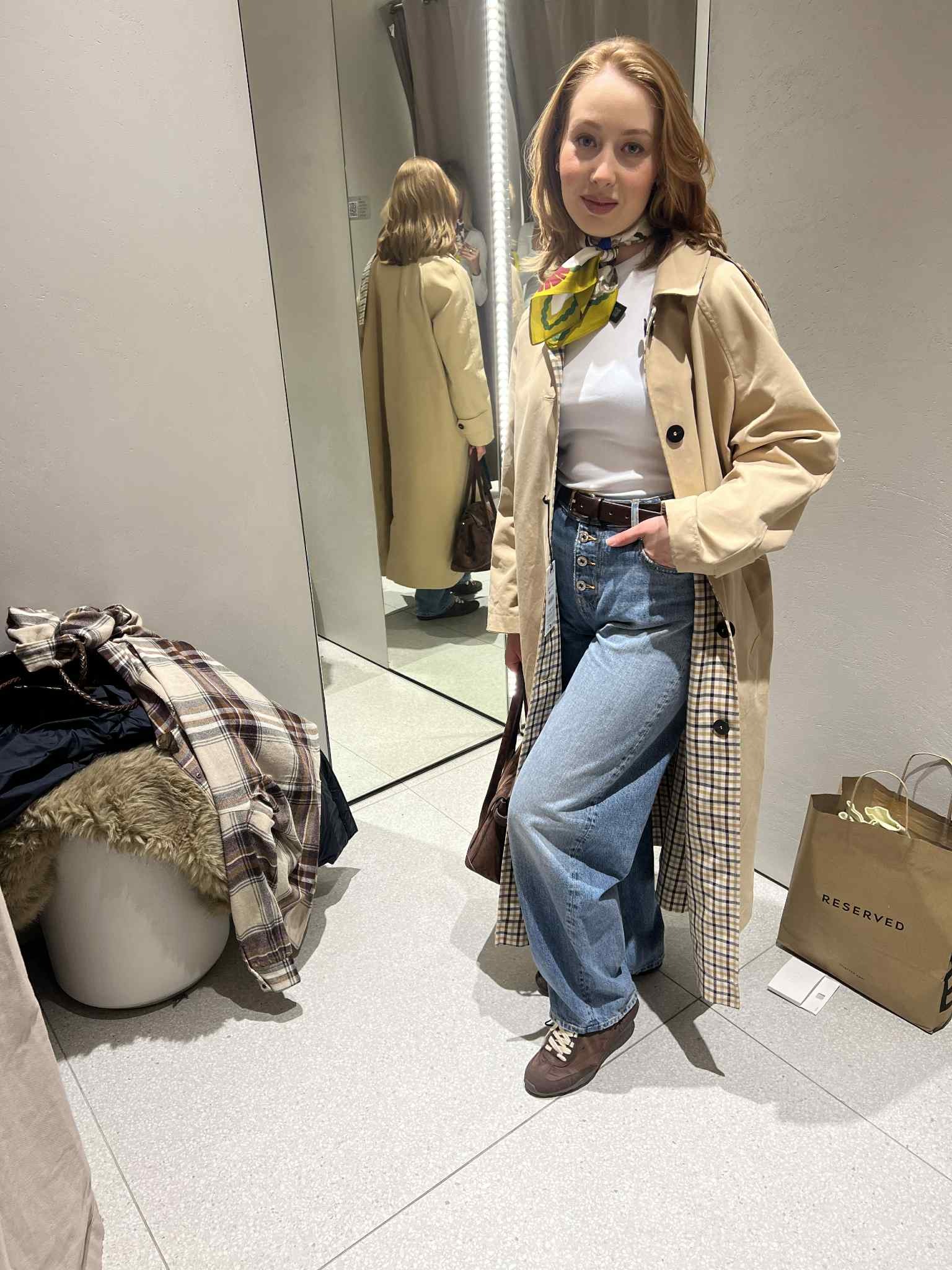 Kobieta w przymierzalni, w beżowym płaszczu, dżinsach i apaszce, pozuje przed lustrem. Widoczne ubrania na pufie i torba Reserved.