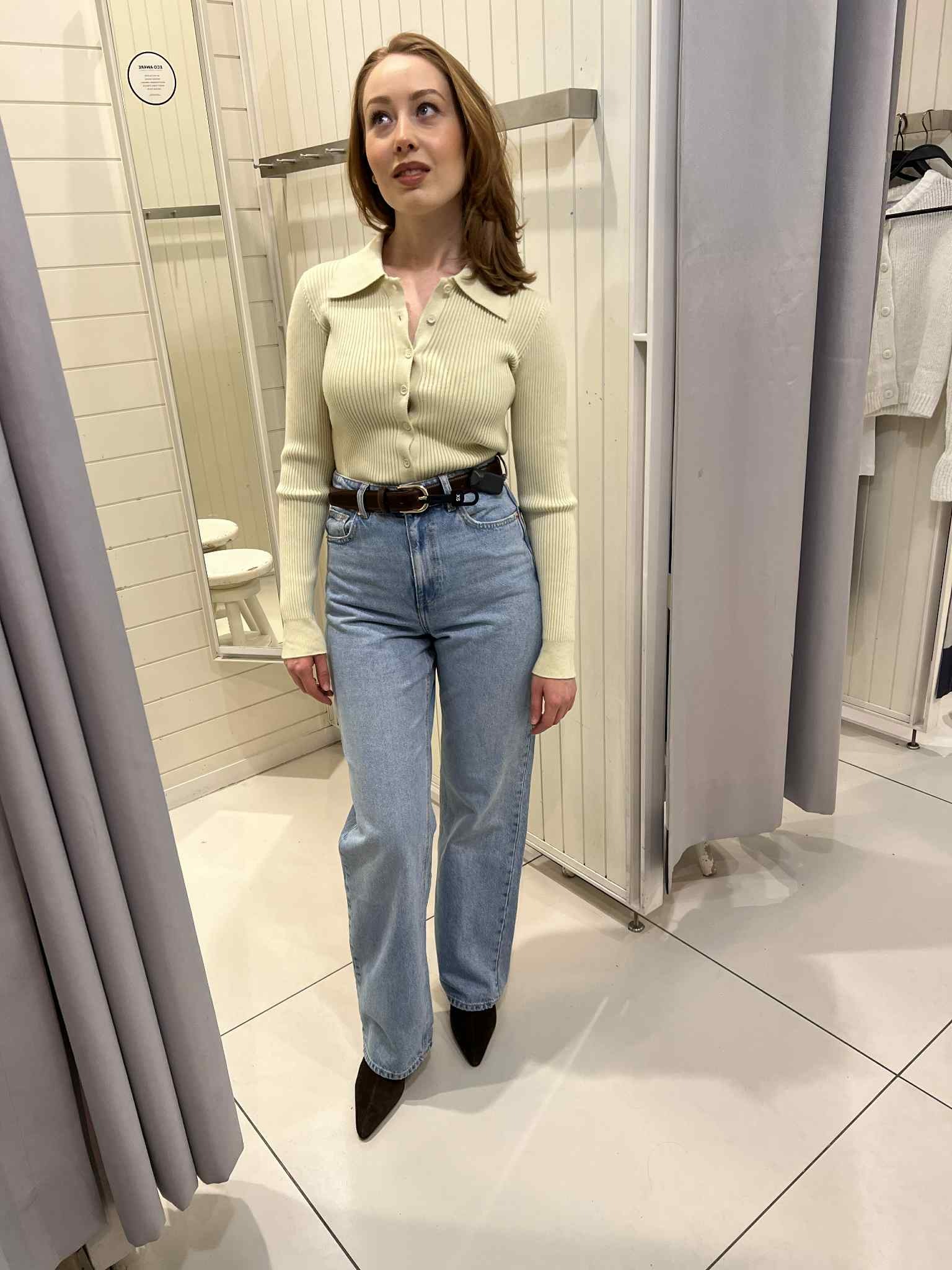 Kobieta w przymierzalni w Warszawie prezentuje stylizację: prążkowany sweter, jeansy z paskiem i szpilki. Neutralne tło, naturalne światło.