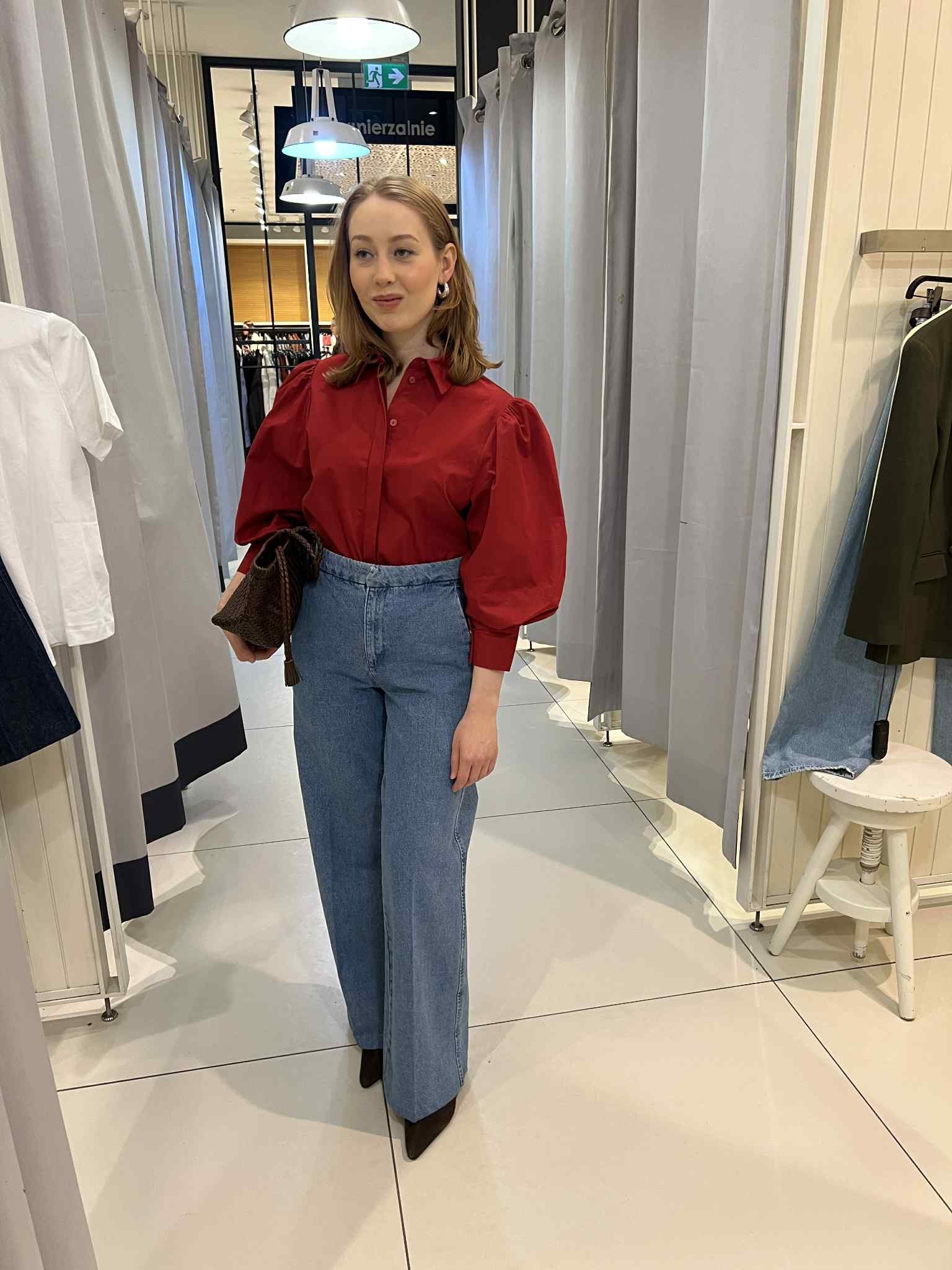 Kobieta w czerwonej bluzce z bufiastymi rękawami i szerokich jeansach, trzymająca torebkę, w przymierzalni z zasłonami i krzesłem.