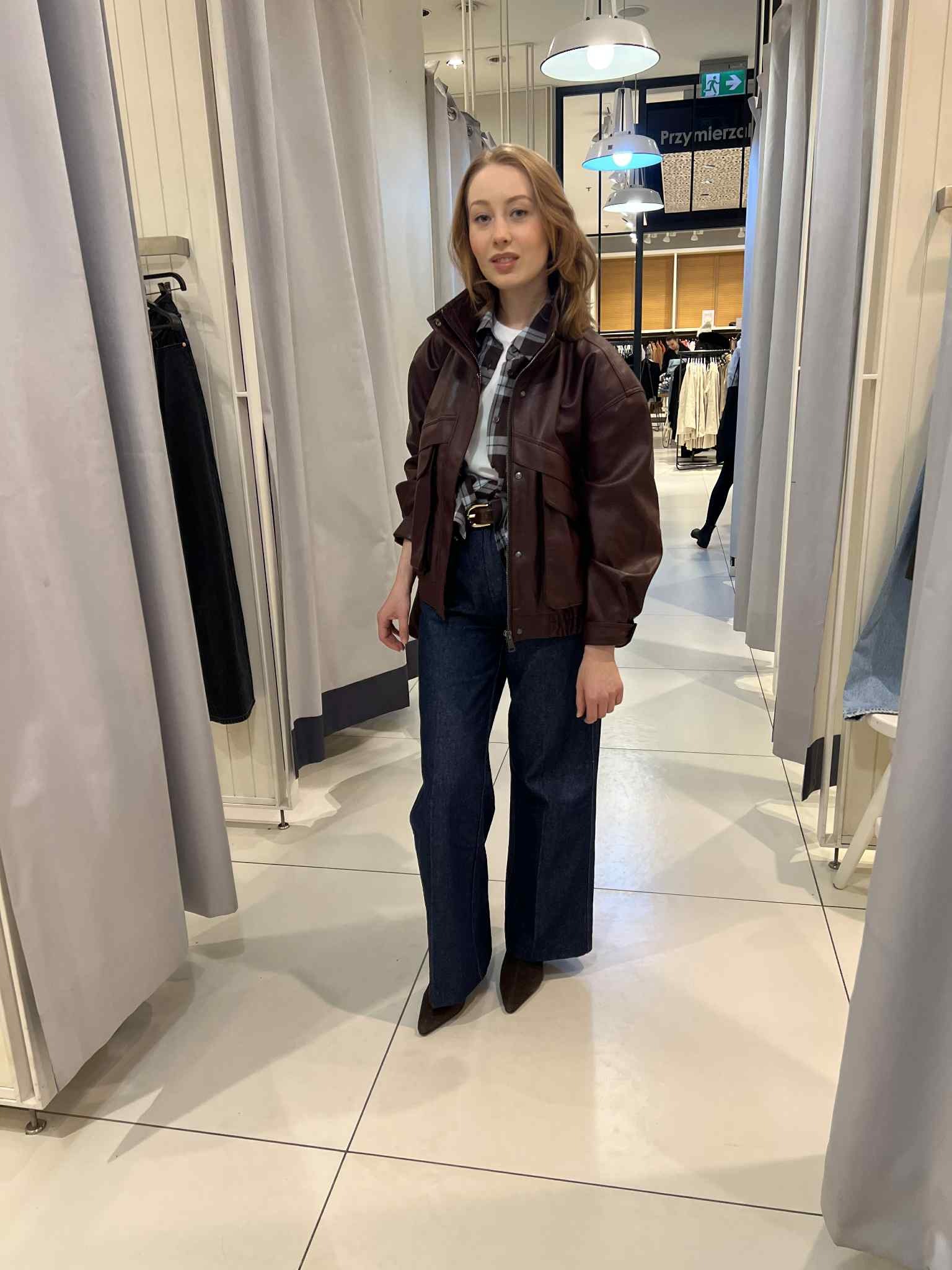 Kobieta w przymierzalni prezentuje stylizację: szerokie jeansy, brązowa kurtka, koszula w kratę i pasek. Widoczne zasłony przymierzalni i oświetlenie.