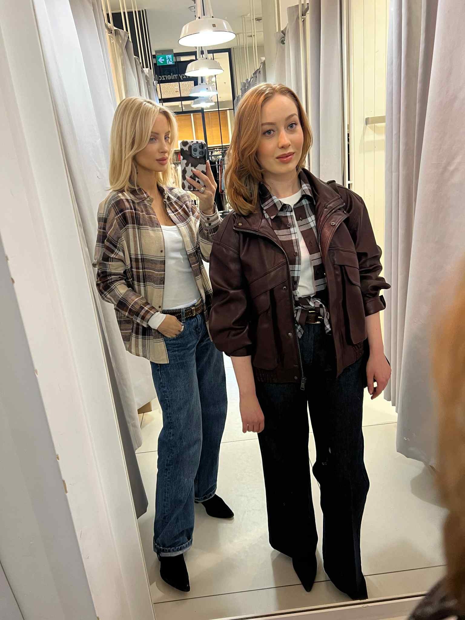Dwie kobiety w przymierzalni, prezentujące różne stylizacje: jedna w kraciastej koszuli i jeansach, druga w skórzanej kurtce i szerokich spodniach. Lustro odbija oświetlenie.