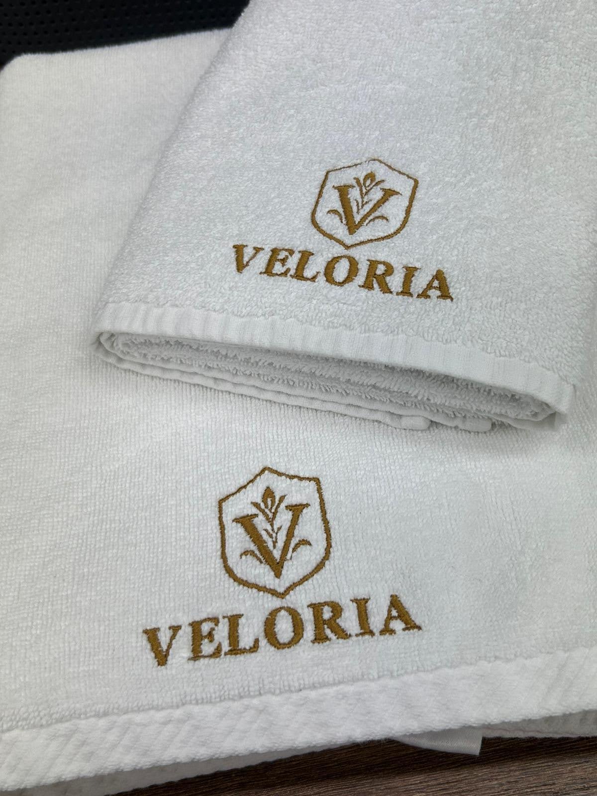Białe ręczniki frotte z wyhaftowanym złotym logo i nazwą 'VELORIA', ułożone jeden na drugim. Widoczne detale haftu i faktura materiału.