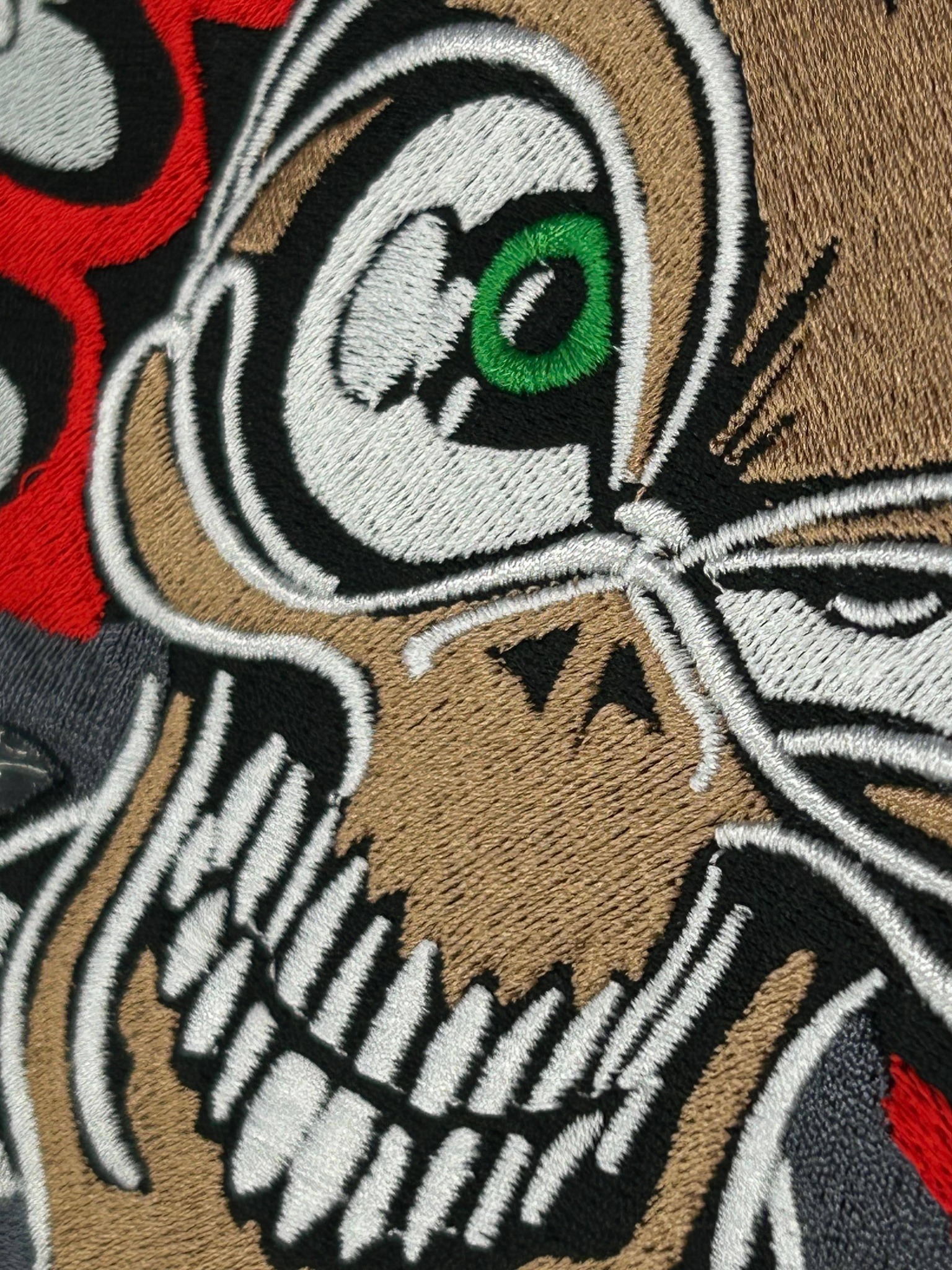 Zbliżenie na haftowaną aplikację z motywem czaszki z zielonym okiem, wykonaną z brązowych, białych, czarnych i czerwonych nici na ciemnym tle.