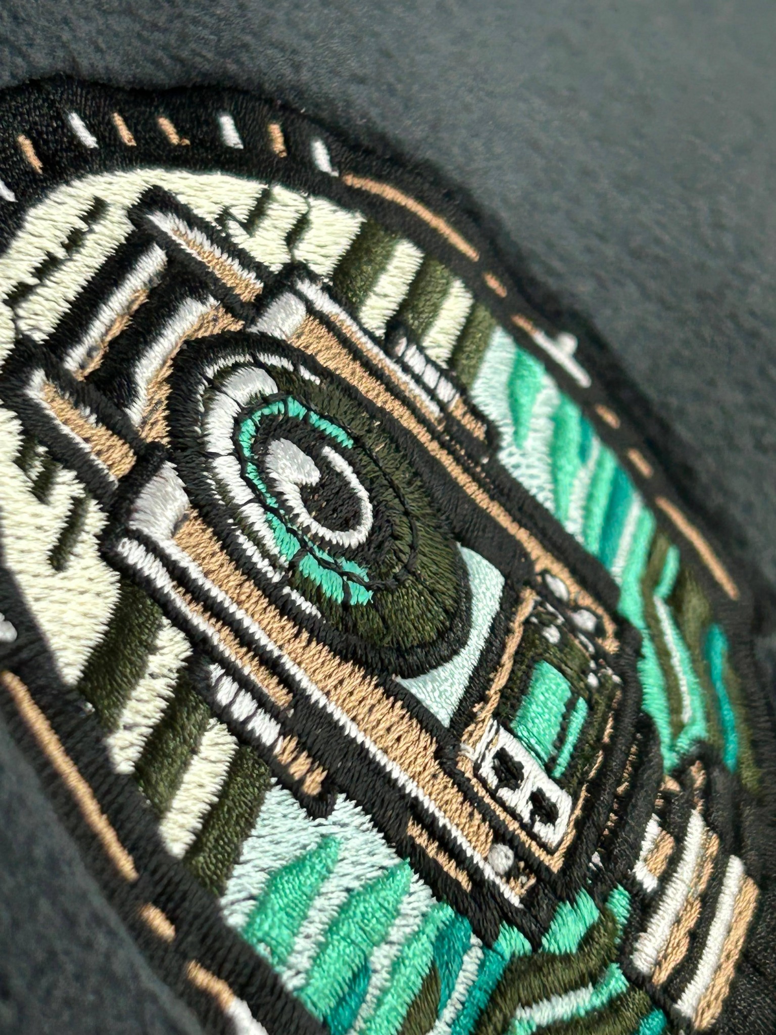 Zbliżenie na haftowany wzór geometryczny w odcieniach zieleni, beżu, czerni i bieli na szarym tle materiału. Widoczne detale ściegów i tekstury haftu.
