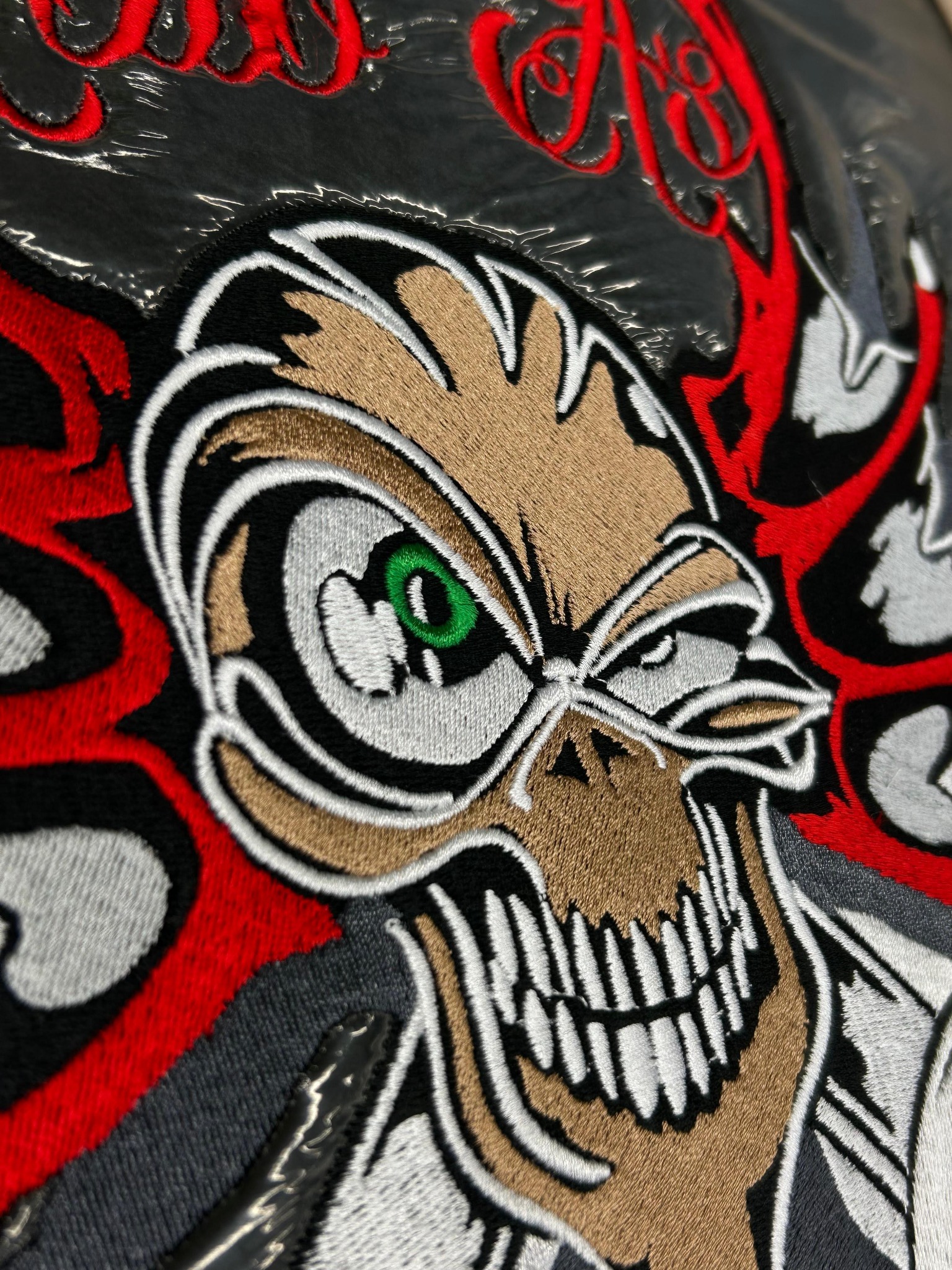 Zbliżenie na haftowany wzór czaszki z zielonym okiem i czerwonymi elementami na czarnym tle, częściowo pokryty folią ochronną. Detaliczne wykonanie.