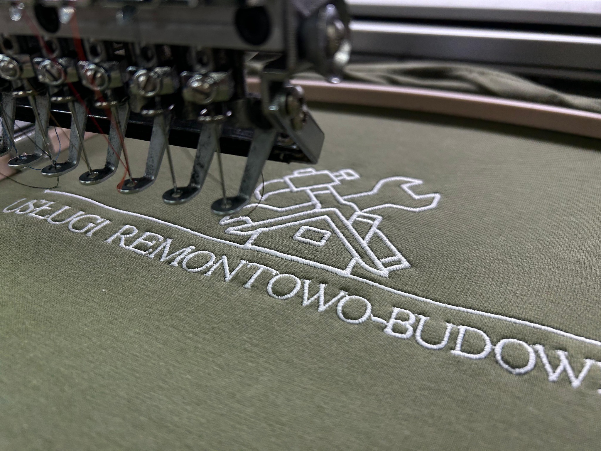 Haft komputerowy na oliwkowej koszulce z logo 'Usługi Remontowo-Budowlane' w trakcie realizacji. Widoczna igła maszyny hafciarskiej i fragment tamborka.