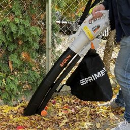 Molly - Osoba obsługuje dmuchawę do liści Prime3 3500W na tle żywopłotu i opadłych liści. Widoczny fragment ogrodzenia i butów operatora.